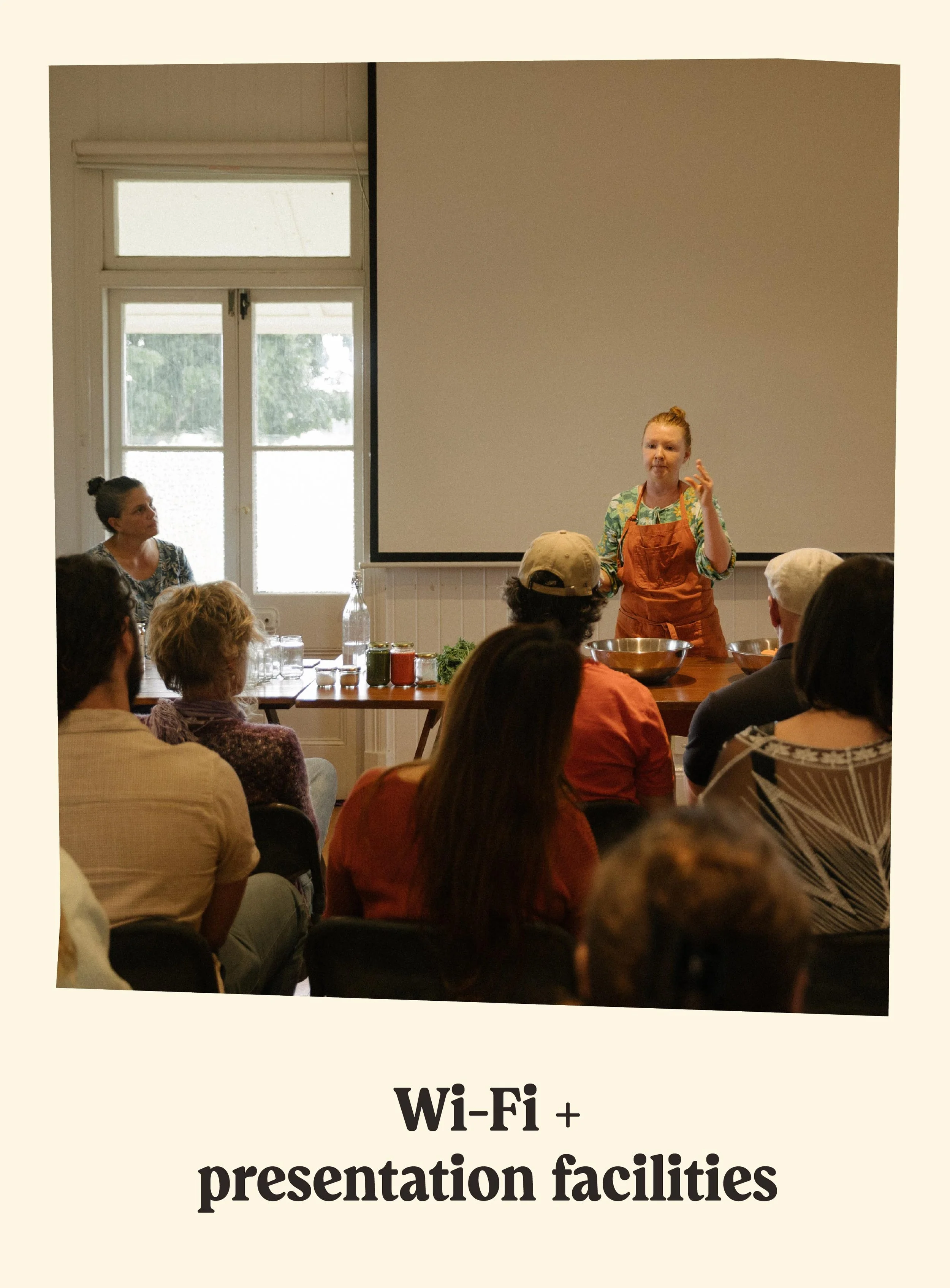 A woman in an orange apron giving a presentation to an audience in a room with large windows and a blank presentation screen.