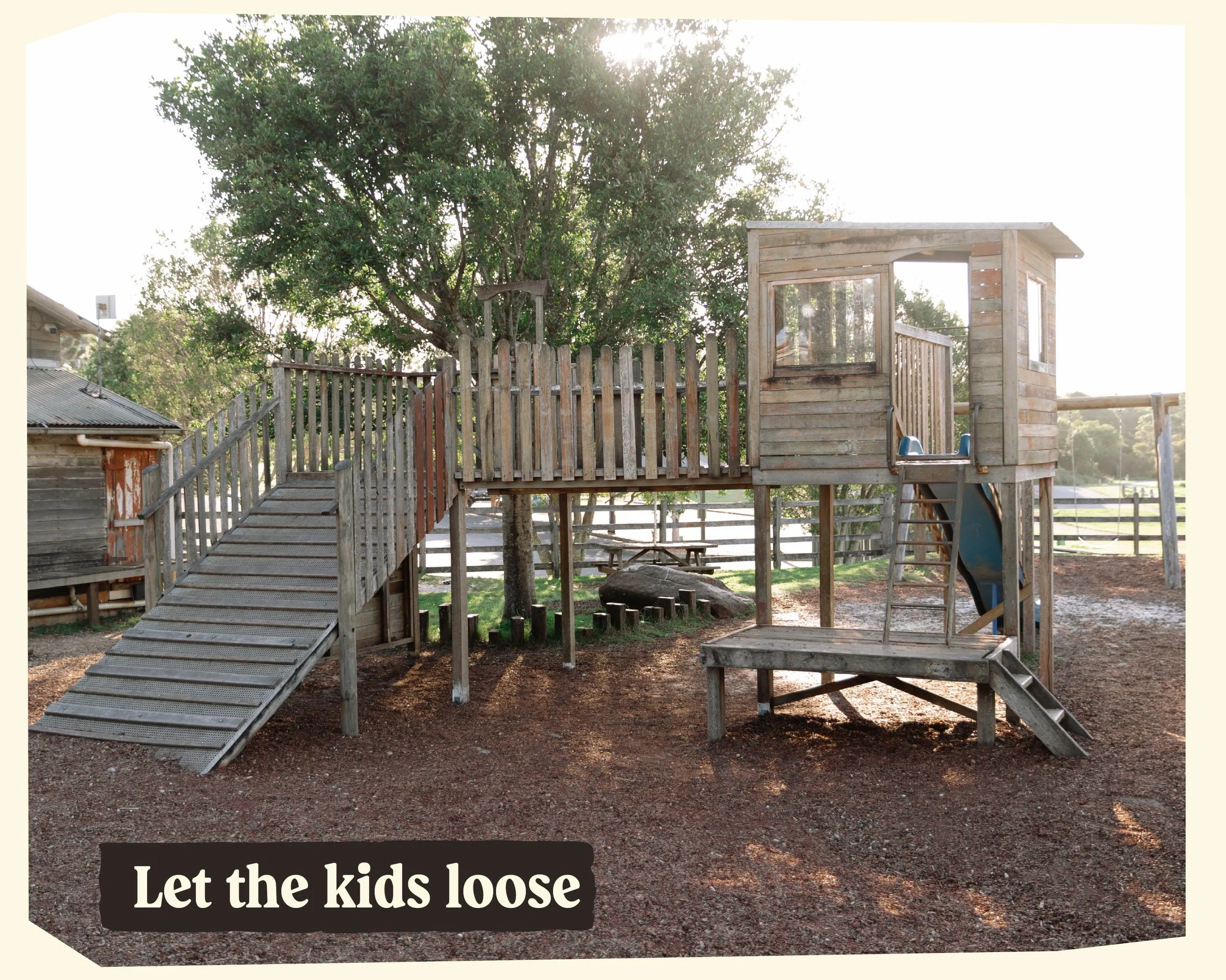 A wooden playground structure with a slide and a small playhouse, surrounded by trees and a fence, with sunlight filtering through the leaves.