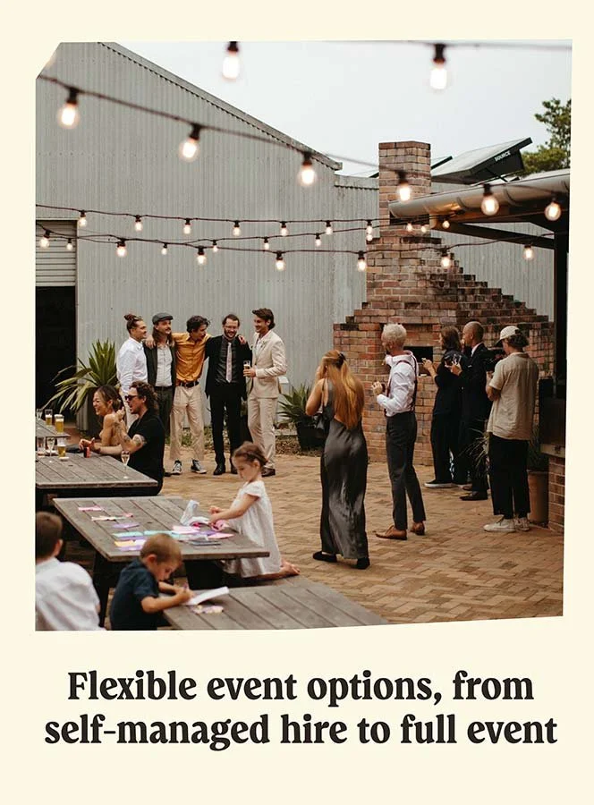 A group of people gathered outdoors for a social event, some standing and talking, others sitting at a table under string lights, with a brick chimney and industrial-style building in the background.