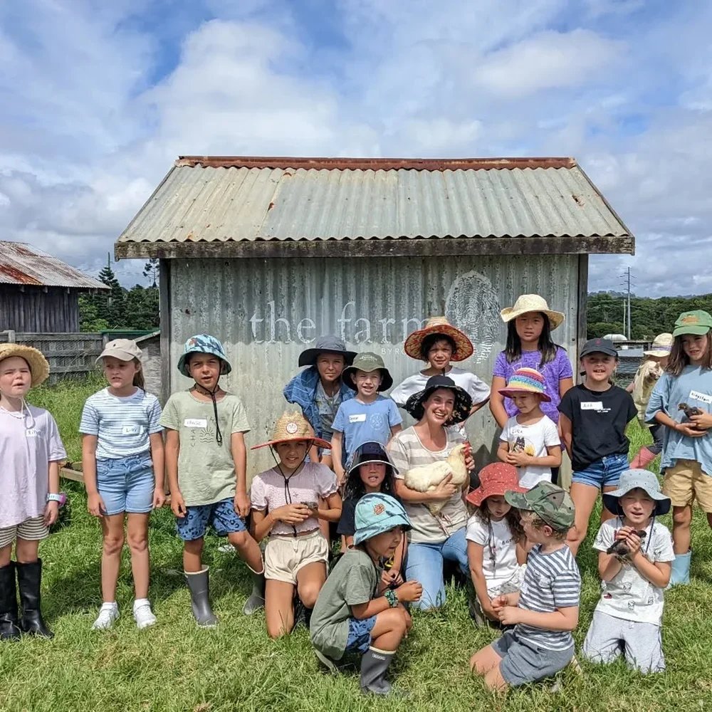 A group of children and an adult woman with a chicken, in front of a weathered farm shed with