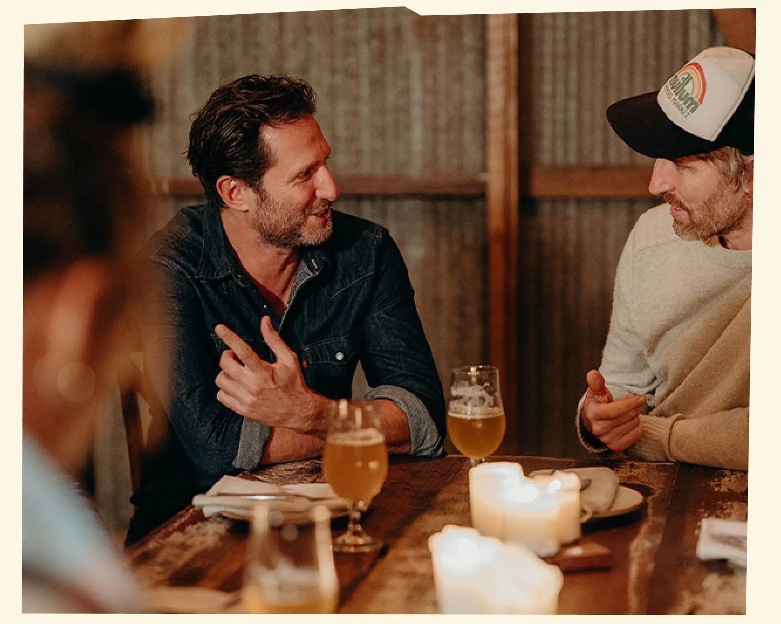 Two men sitting at a bar, engaging in conversation with drinks and candles on the table, in a cozy, dimly-lit setting.