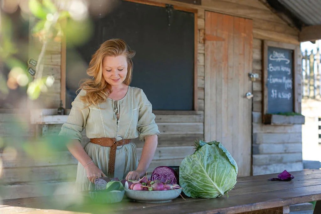 A woman with red hair in a light-colored dress chopping vegetables outdoors at a rustic wooden stand, with a large cabbage and other vegetables on the table.