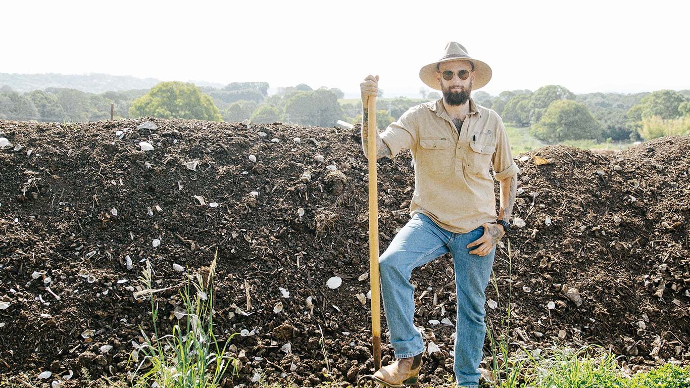 A man with a beard wearing sunglasses, a wide-brimmed hat, a beige shirt, and jeans, standing outdoors beside a mound of dirt, holding a shovel, with a background of trees and greenery.