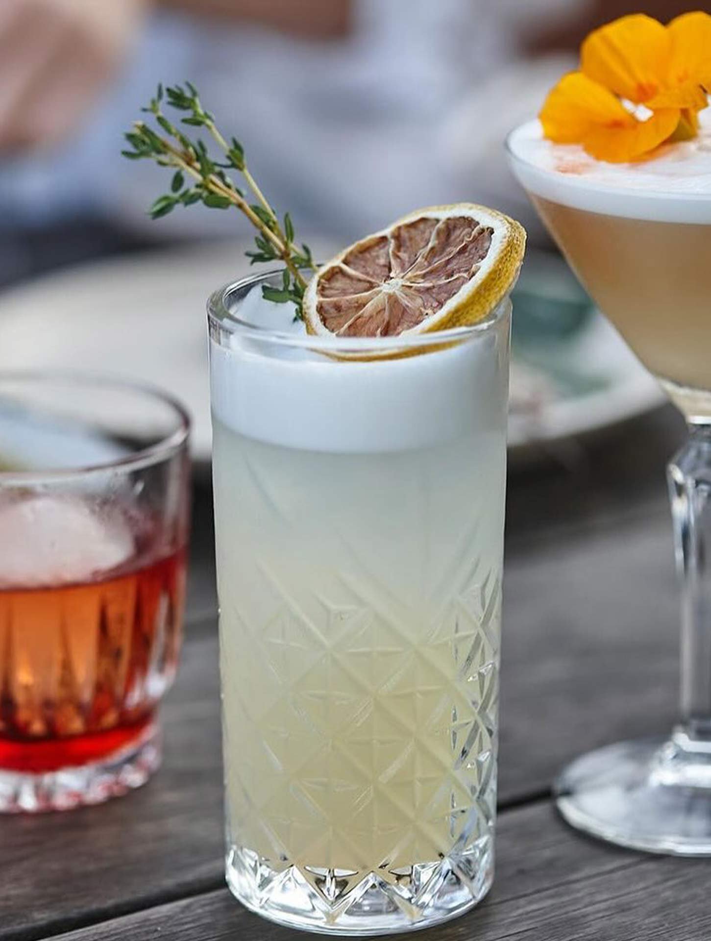 A tall glass containing a pale yellow-green beverage garnished with a dried lemon slice and a sprig of thyme, placed on a wooden surface.