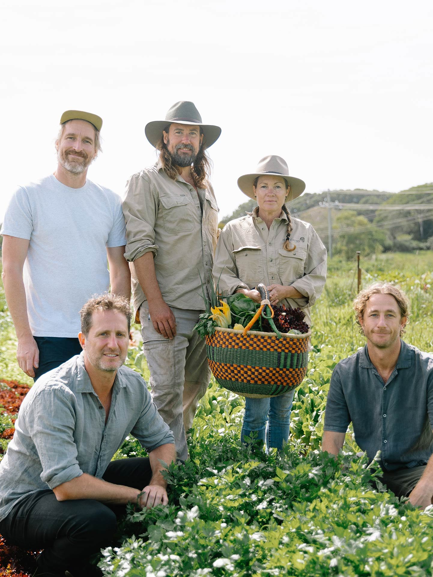 Group of people outdoors in a lush green farm or garden setting, with some holding a basket of fresh produce and vegetables.