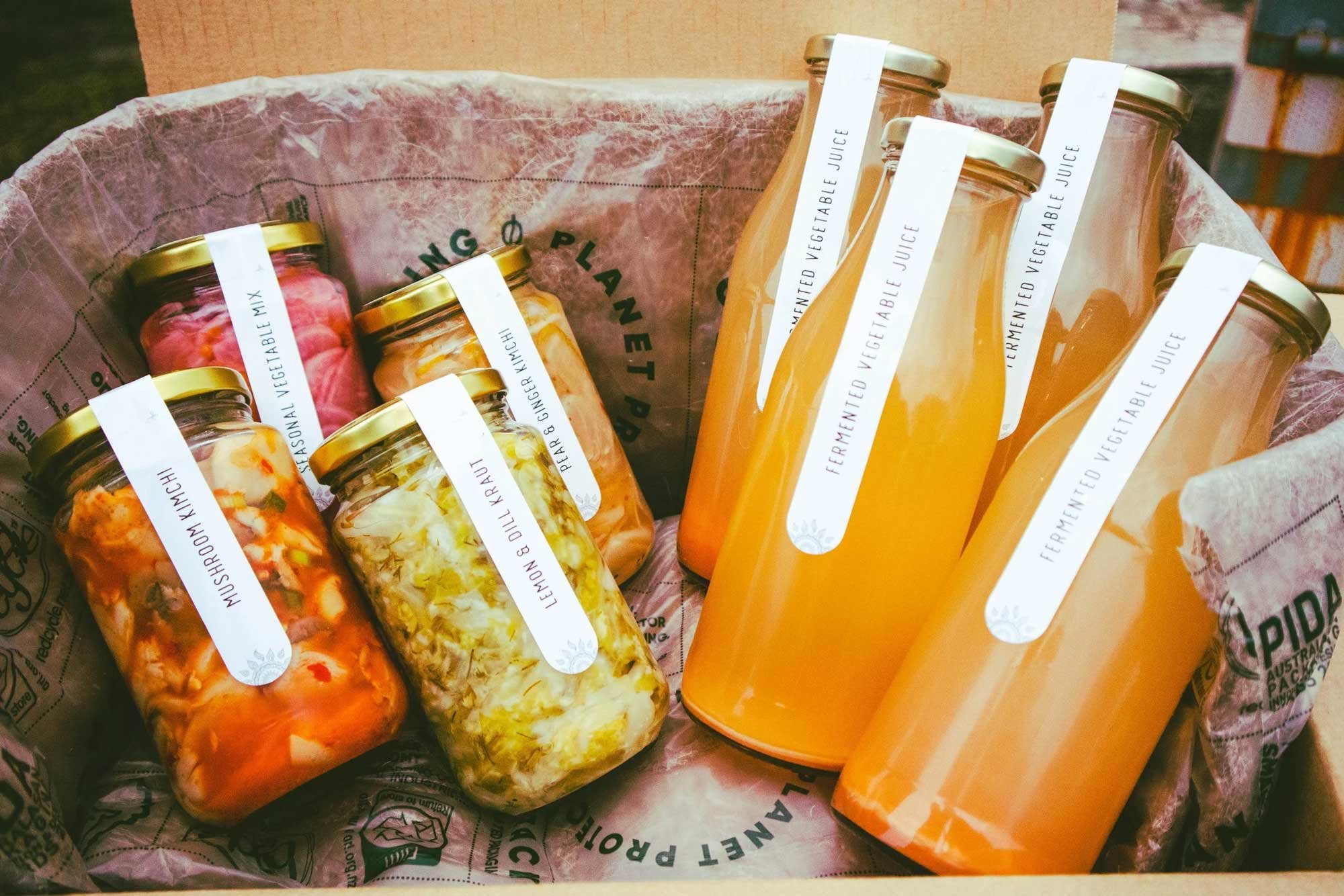 Several glass jars filled with fermented vegetable juice, such as kimchi and sauerkraut, with white labels and gold lids, placed inside a paper-lined basket.
