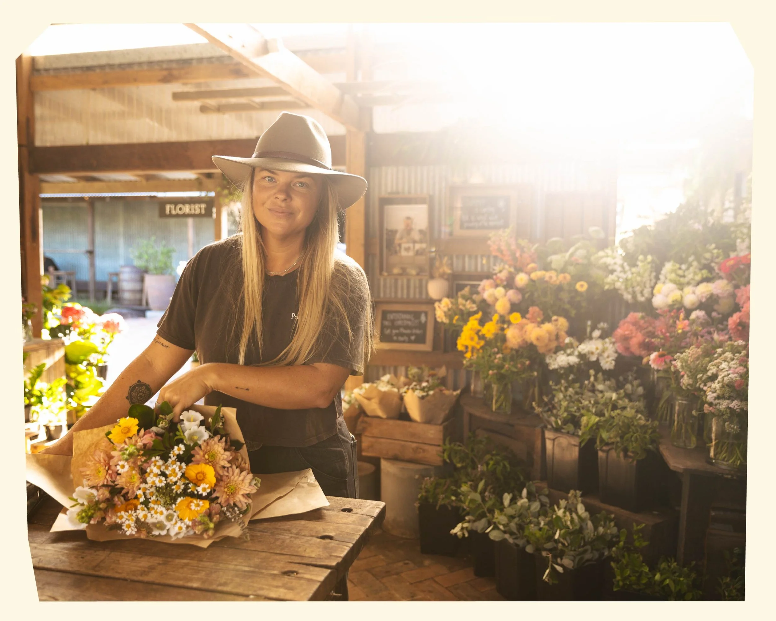 A woman wearing a wide-brimmed hat standing behind a table with a bouquet of colorful flowers at a flower shop. Shelves of flowers and plants are in the background.