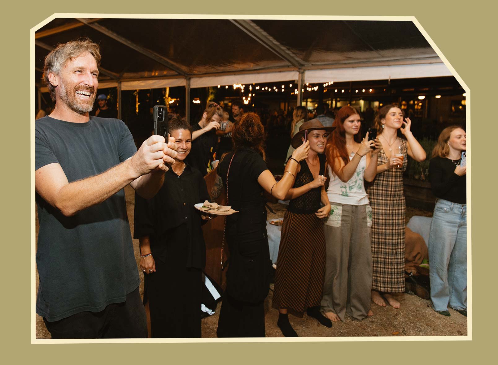 Group of people at an outdoor gathering, smiling and taking photos during the night under a tent with string lights.