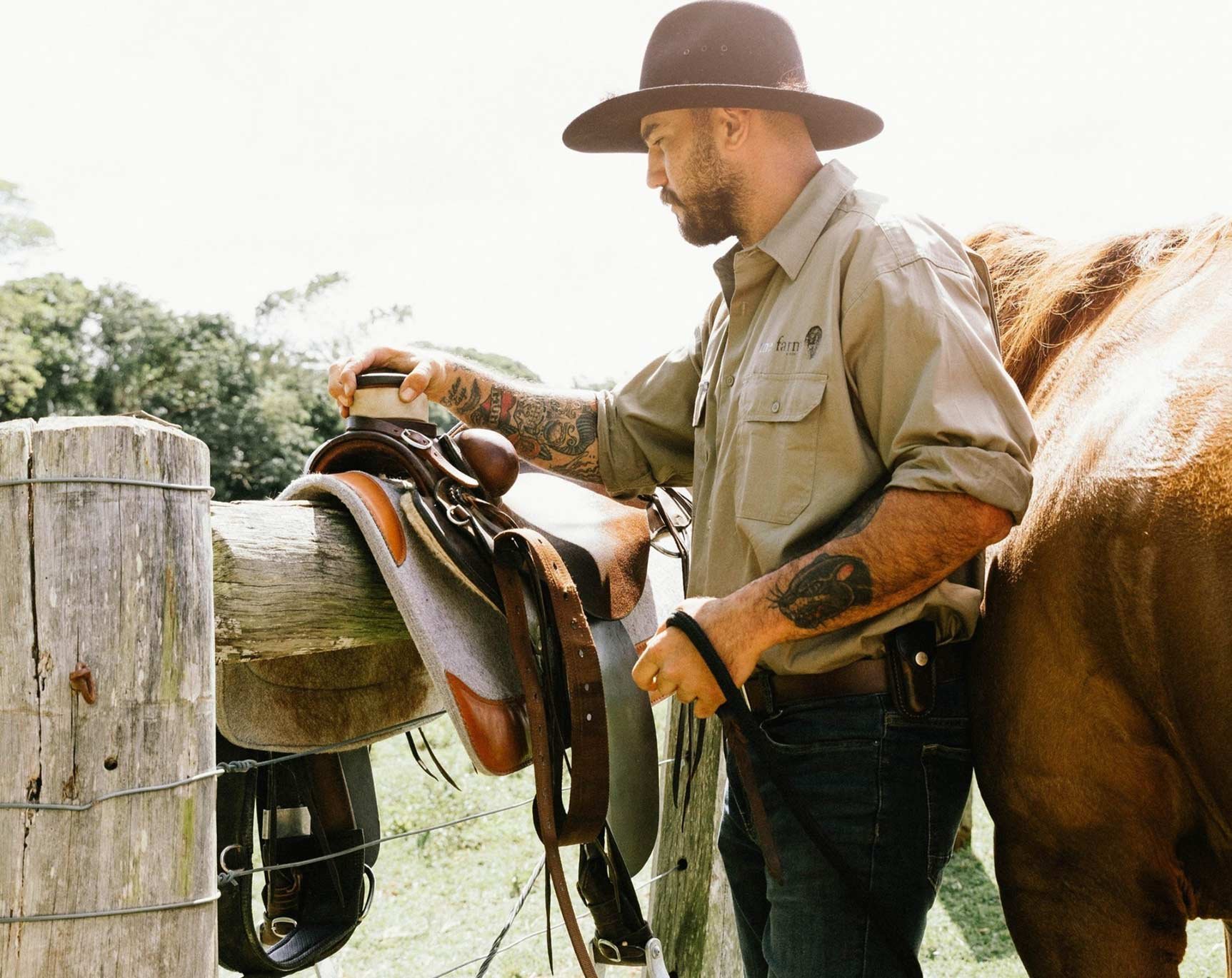 A man with a beard and tattoos wearing a wide-brimmed hat and khaki shirt standing next to a horse, adjusting a saddle on a wooden fence post during daytime.
