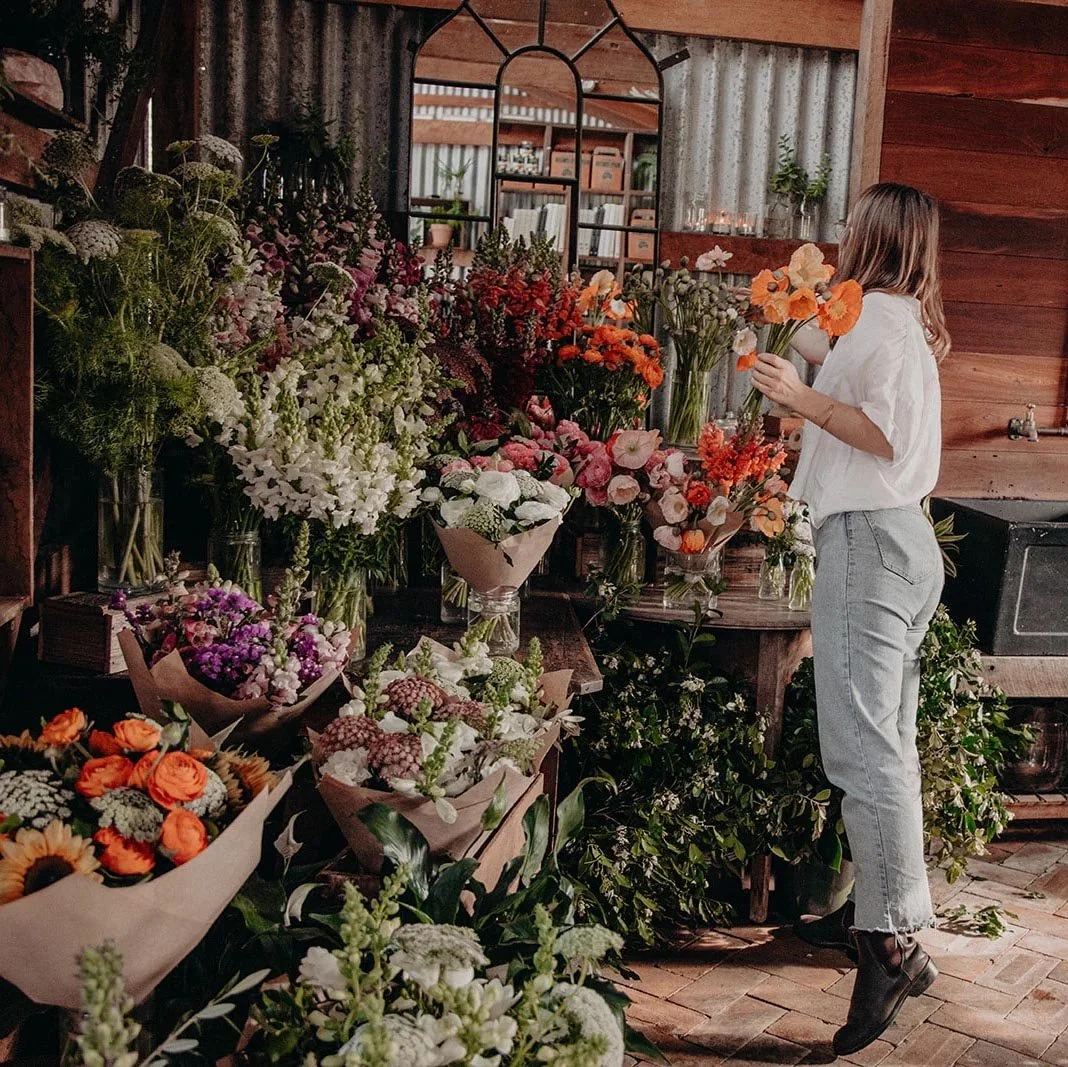 A woman arranging orange flowers in a flower shop with various colorful blooms including roses, peonies, and wildflowers on display.