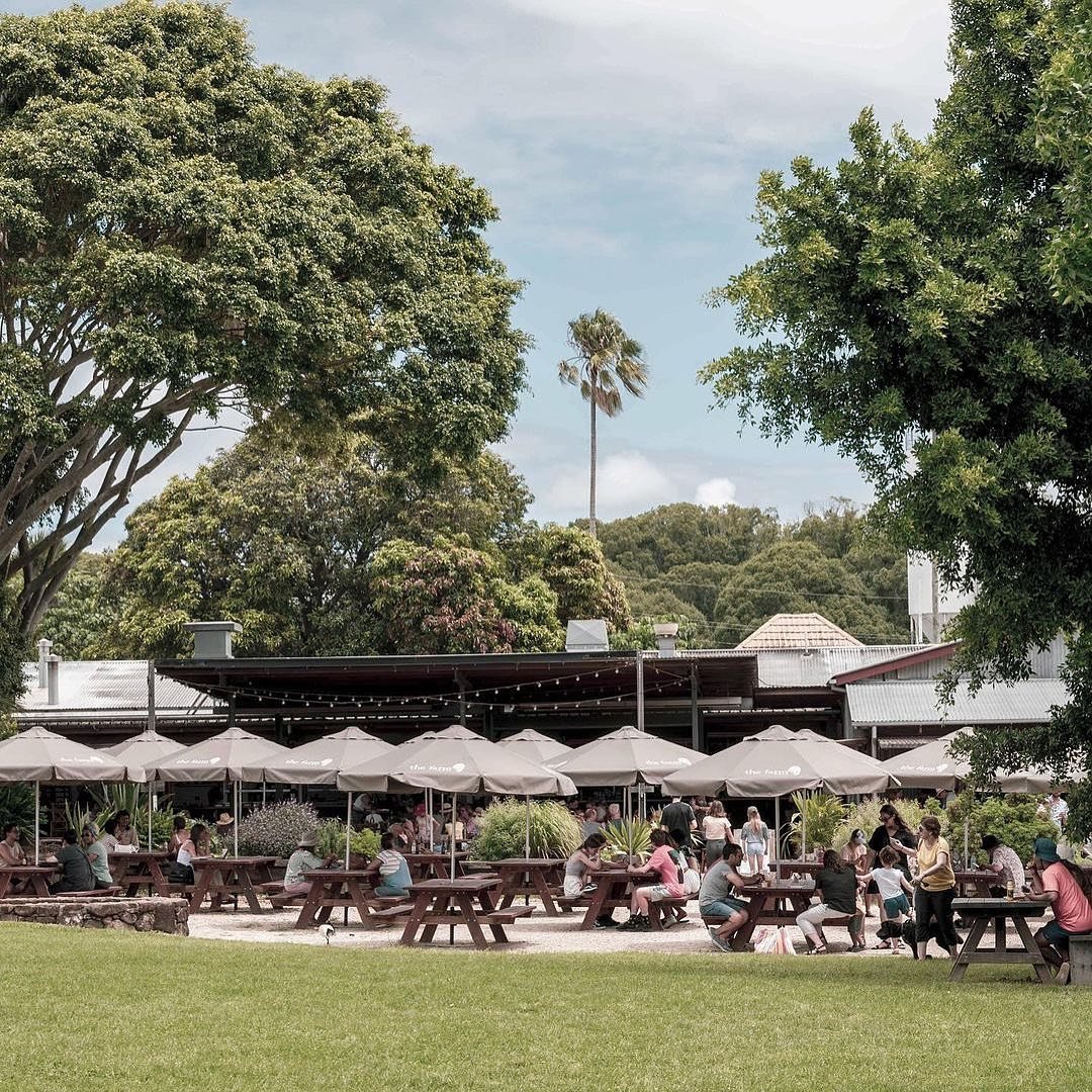 People sitting at outdoor picnic tables under umbrellas at a park or patio area, with large trees and a cloudy sky in the background.