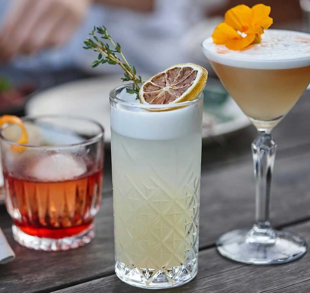 Three cocktails on a wooden table with garnishes, including a lemon slice and herbs.
