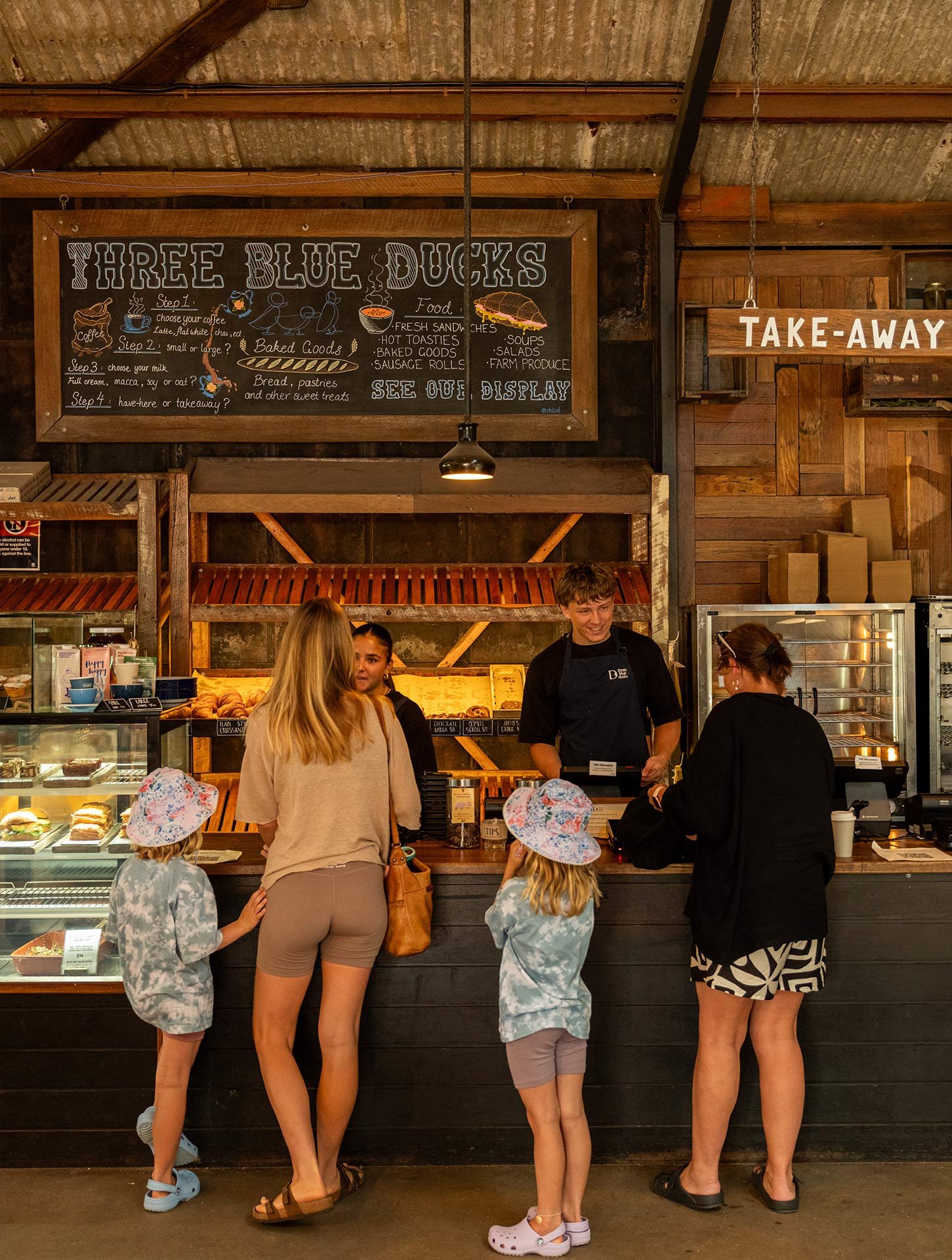 People ordering food at counter in a rustic-style cafe with a chalkboard menu for Three Blue Ducks including options like sandwiches, baked goods, and soups.