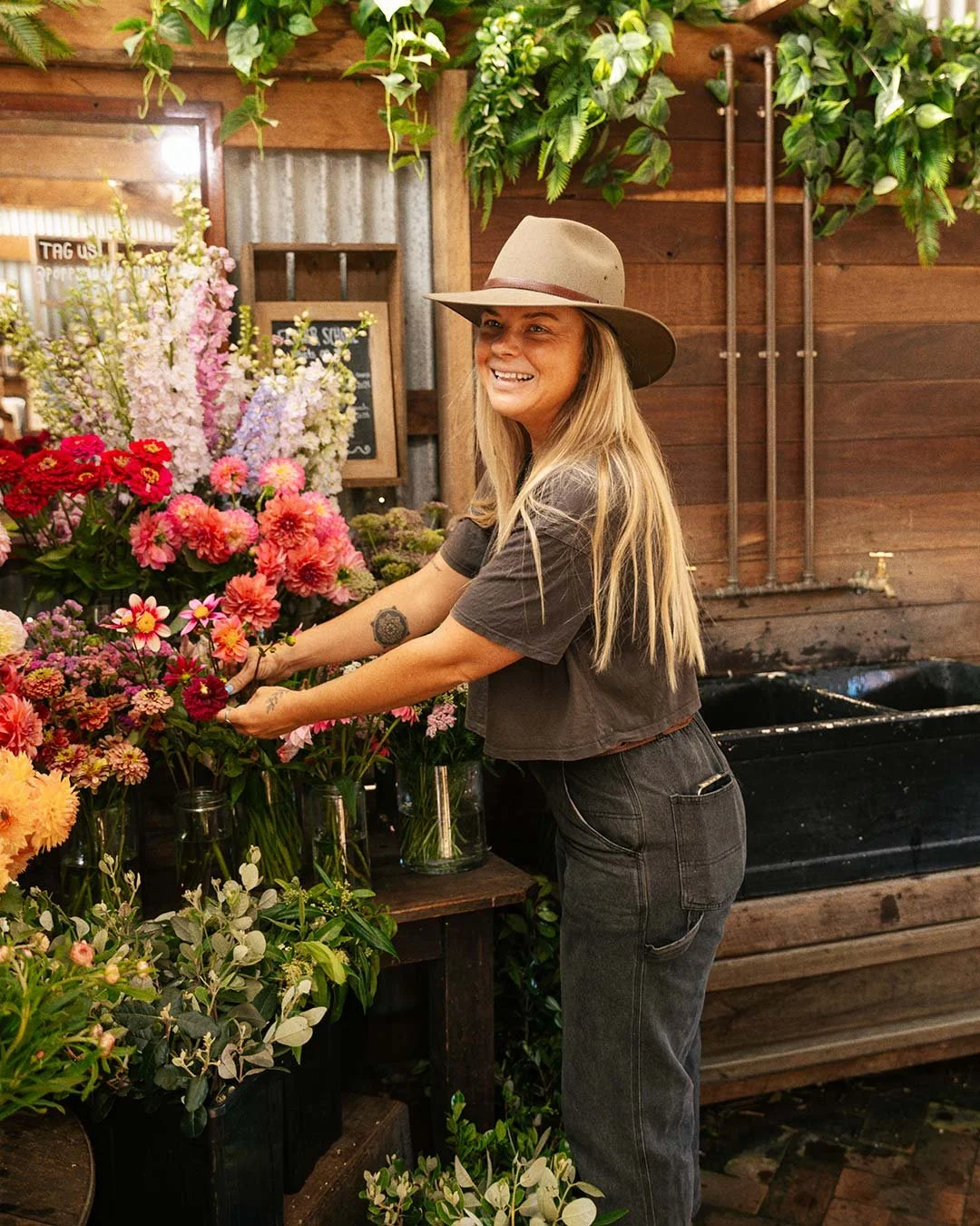 A woman with long blonde hair wearing a beige wide-brimmed hat, a black t-shirt, and black jeans arranging pink, purple, and red flowers in glass vases inside a rustic flower shop decorated with green plants and wooden walls.