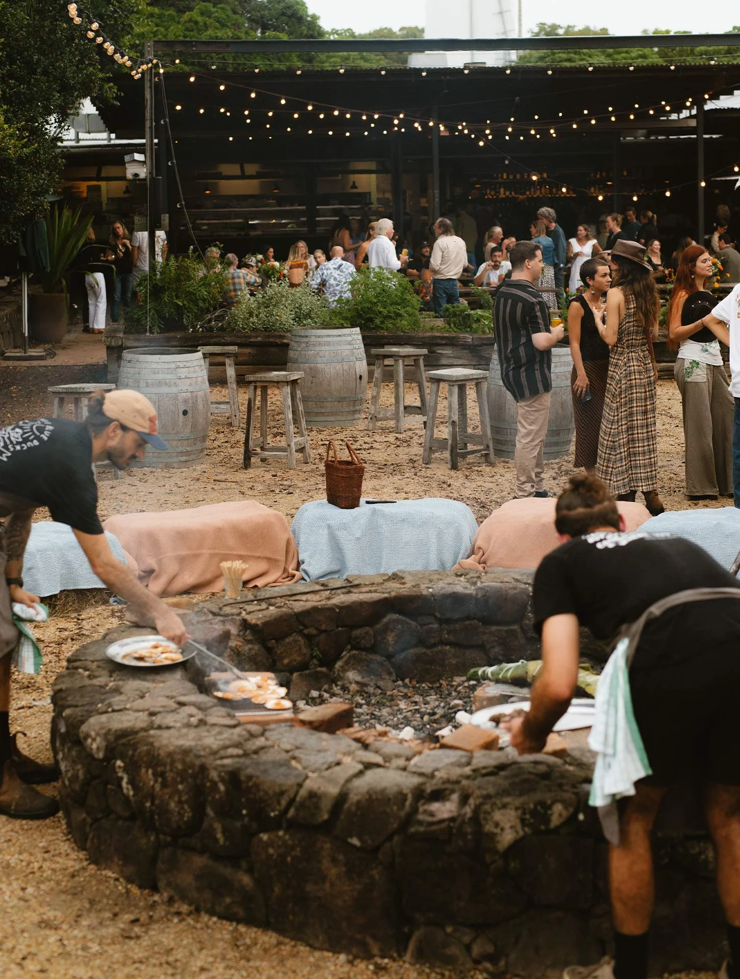 People gathering at an outdoor venue with string lights overhead, some talking and others preparing food over a stone firepit.