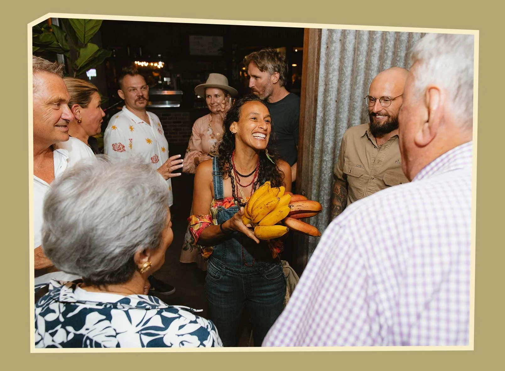 A woman with dark, curly hair wearing denim overalls and colorful clothing, holding a bunch of bananas and talking to a group of people at a social gathering in a restaurant or bar.