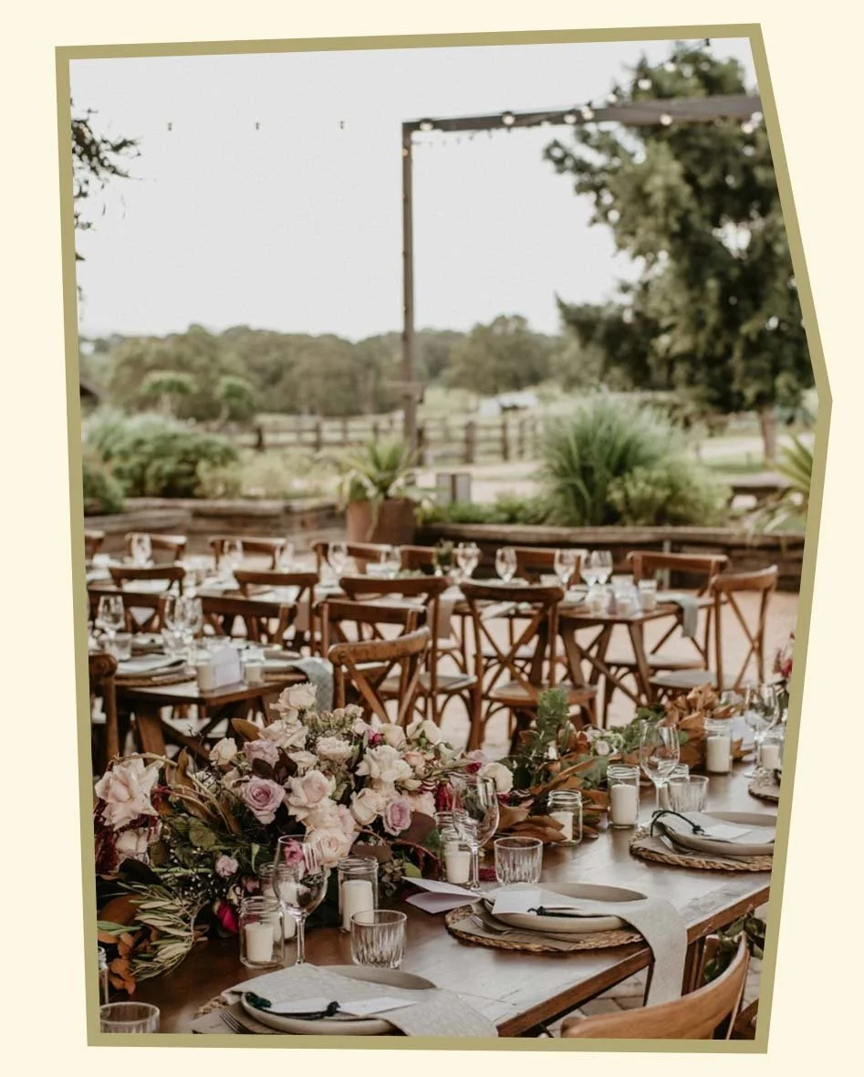 Decorated outdoor dining table with flowers, candles, and tableware at a rustic event venue.