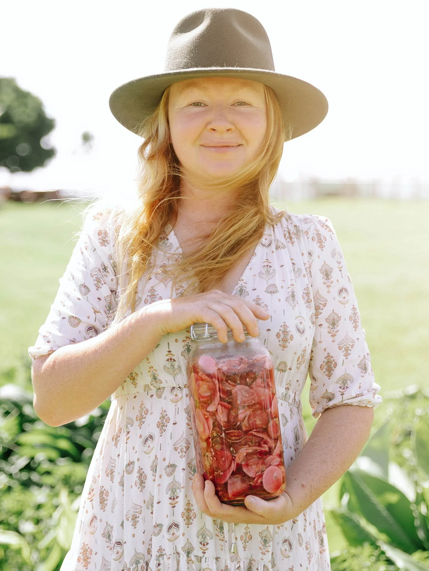 A woman with red hair holding a glass jar filled with sliced strawberries and raspberries, outdoors in a bright, sunny farm or garden setting.