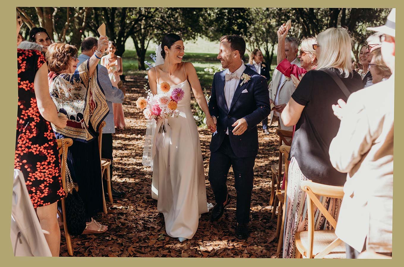 A newlywed couple smiling and holding hands in an outdoor wedding, surrounded by guests clapping and celebrating under trees with a leafy ground.
