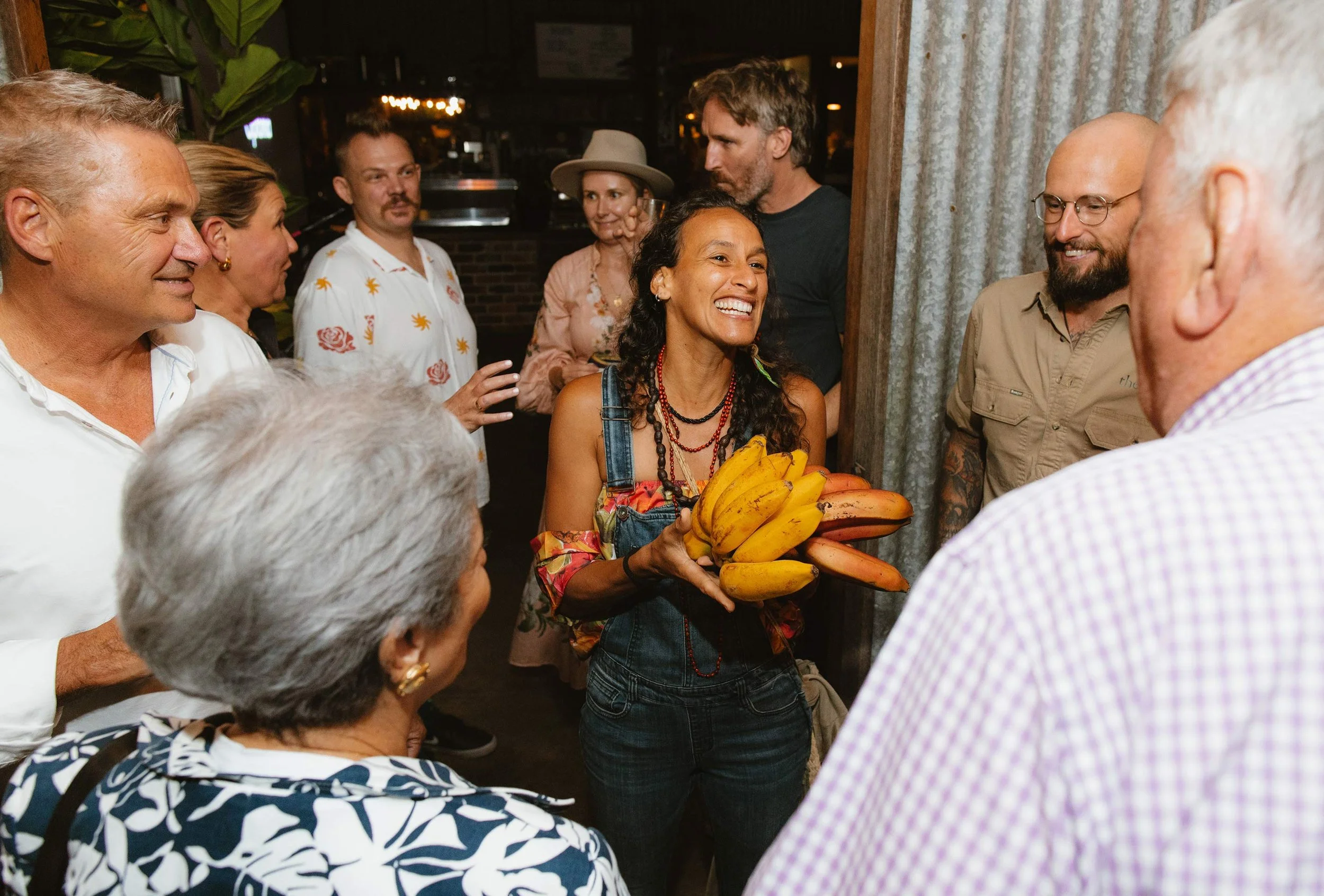 Woman holding a bunch of bananas, smiling and surrounded by a group of people at a social gathering.