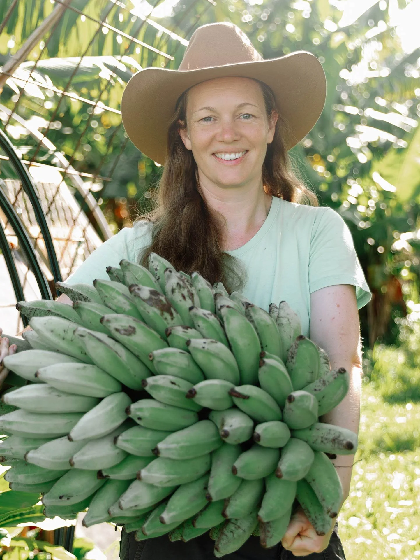 A woman wearing a large hat smiling while holding a bunch of green bananas in a lush, green outdoor setting.