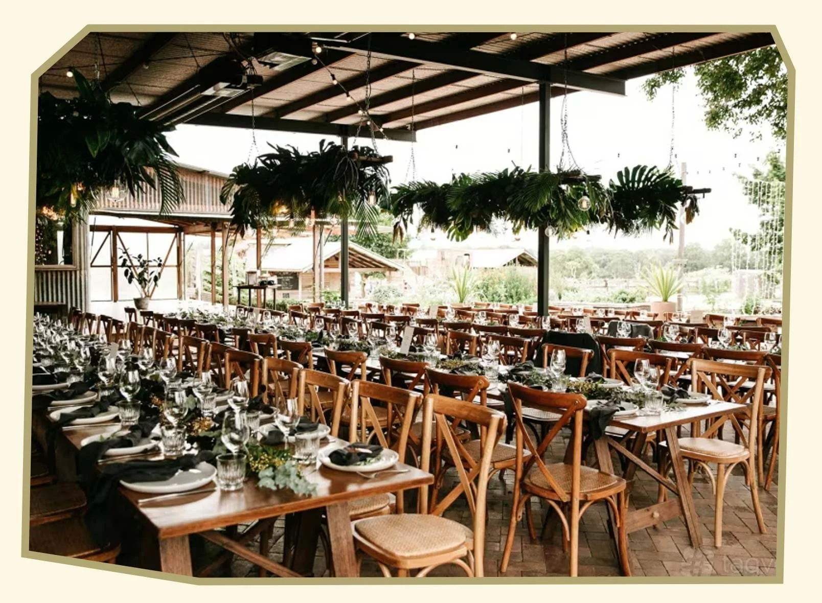 Indoor reception area set up for a wedding or event with long wooden tables decorated with black napkins, glassware, and floral centerpieces, overlooking an outdoor garden view.