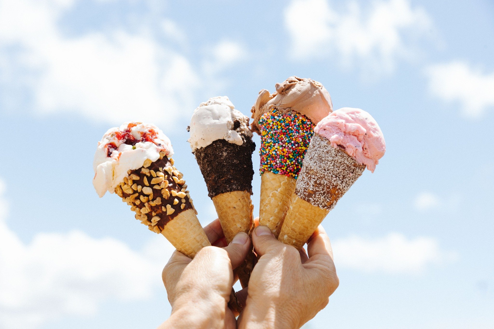 Hand holding five ice cream cones with various flavors and toppings against a cloudy sky