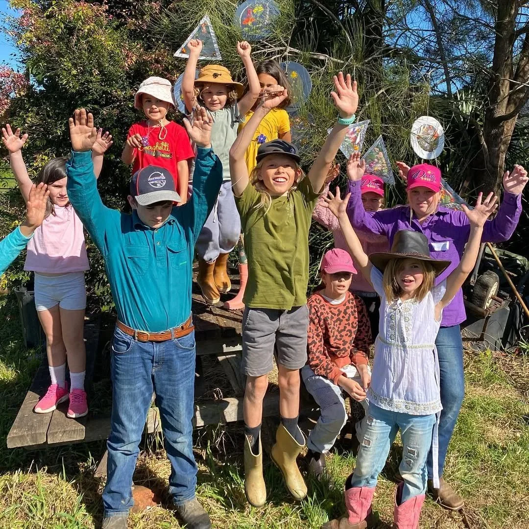 Group of children outdoors, smiling and raising their hands in the air, some wearing hats and rain boots, with greenery and trees in the background.