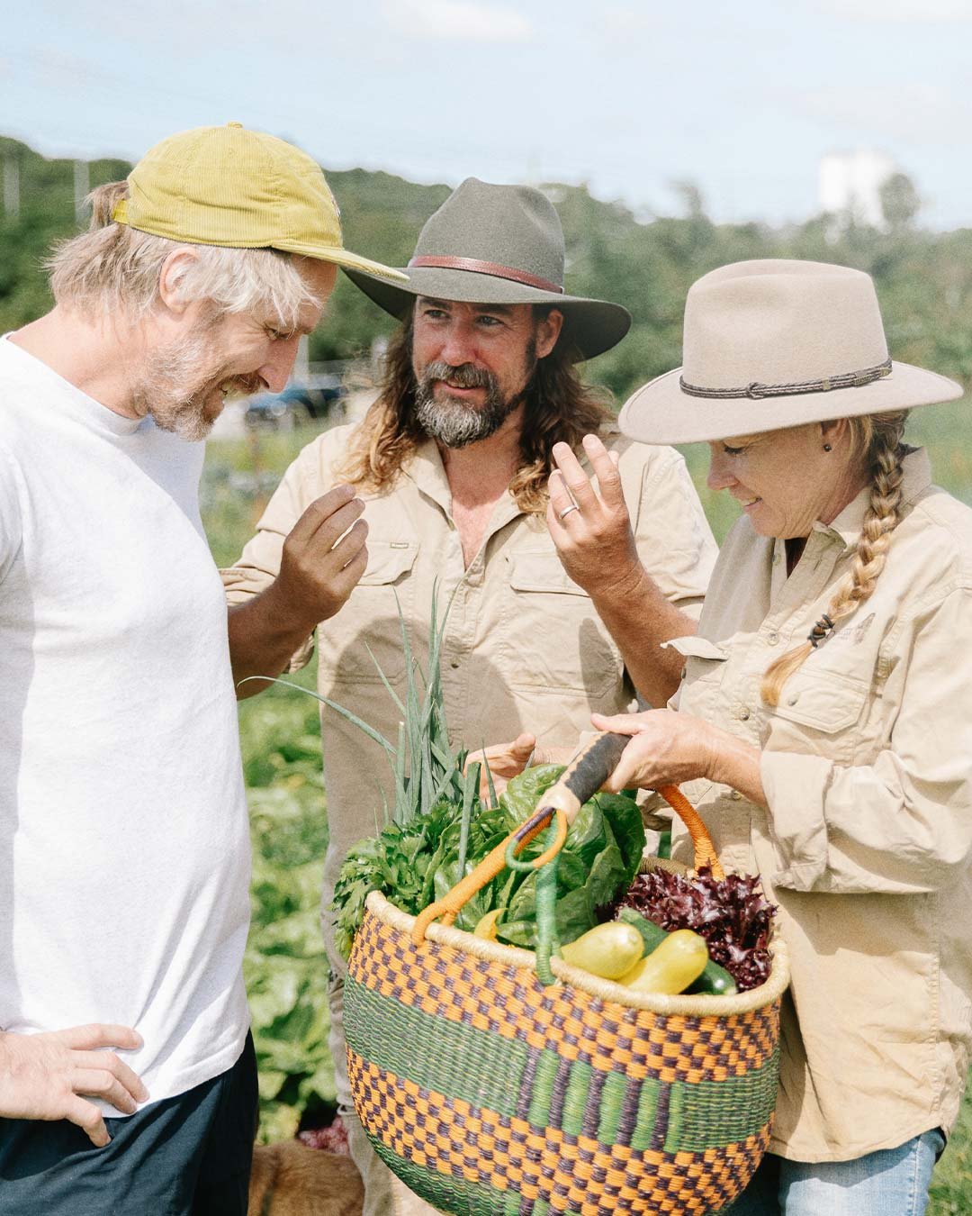 Three people in a garden or farm talking, one woman holding a basket full of vegetables, and the man on the right holding gardening scissors.