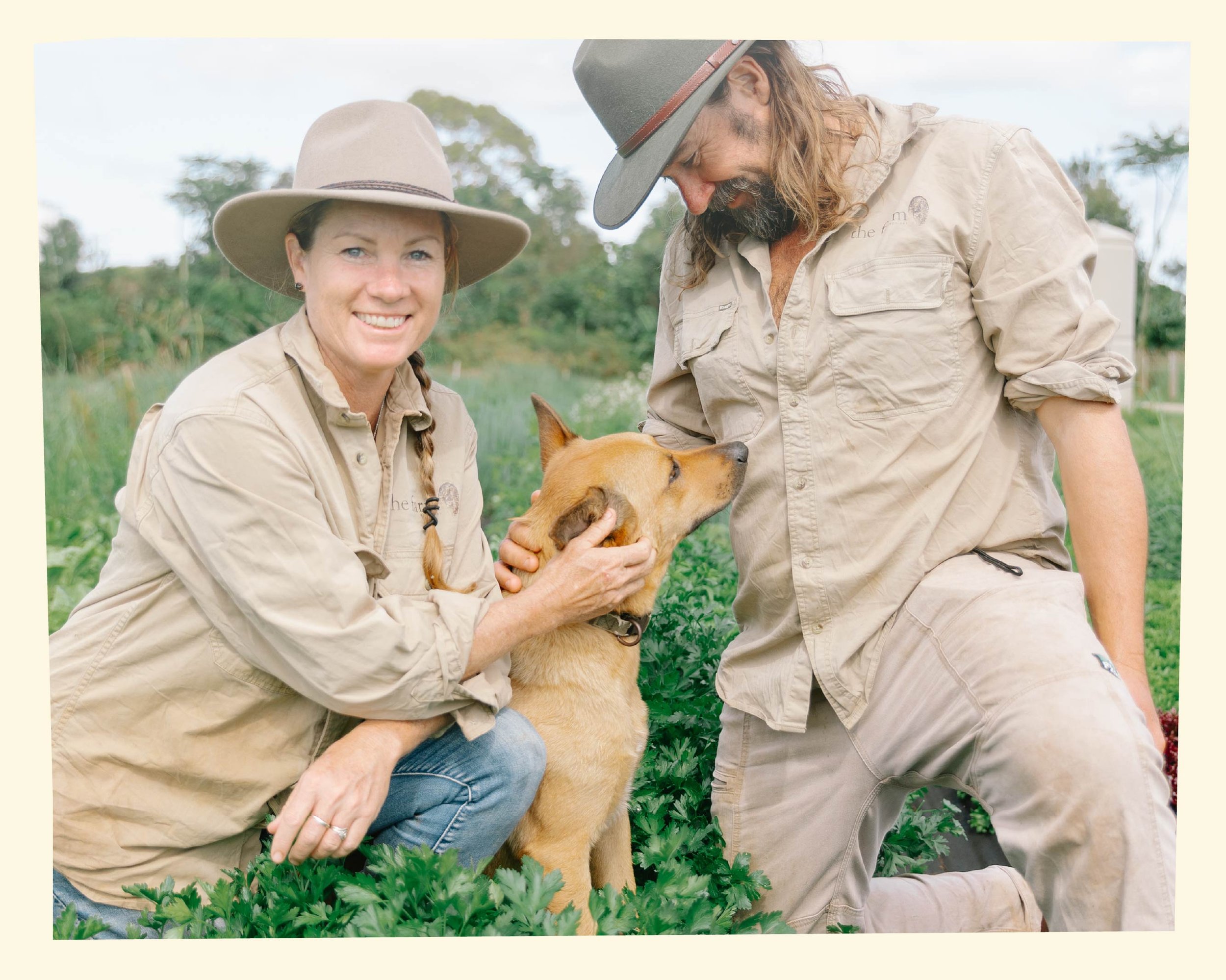 Two people, one woman and one man, are outside in a garden or farm setting with green plants and trees in the background, interacting with a brown dog that they are both holding and petting.