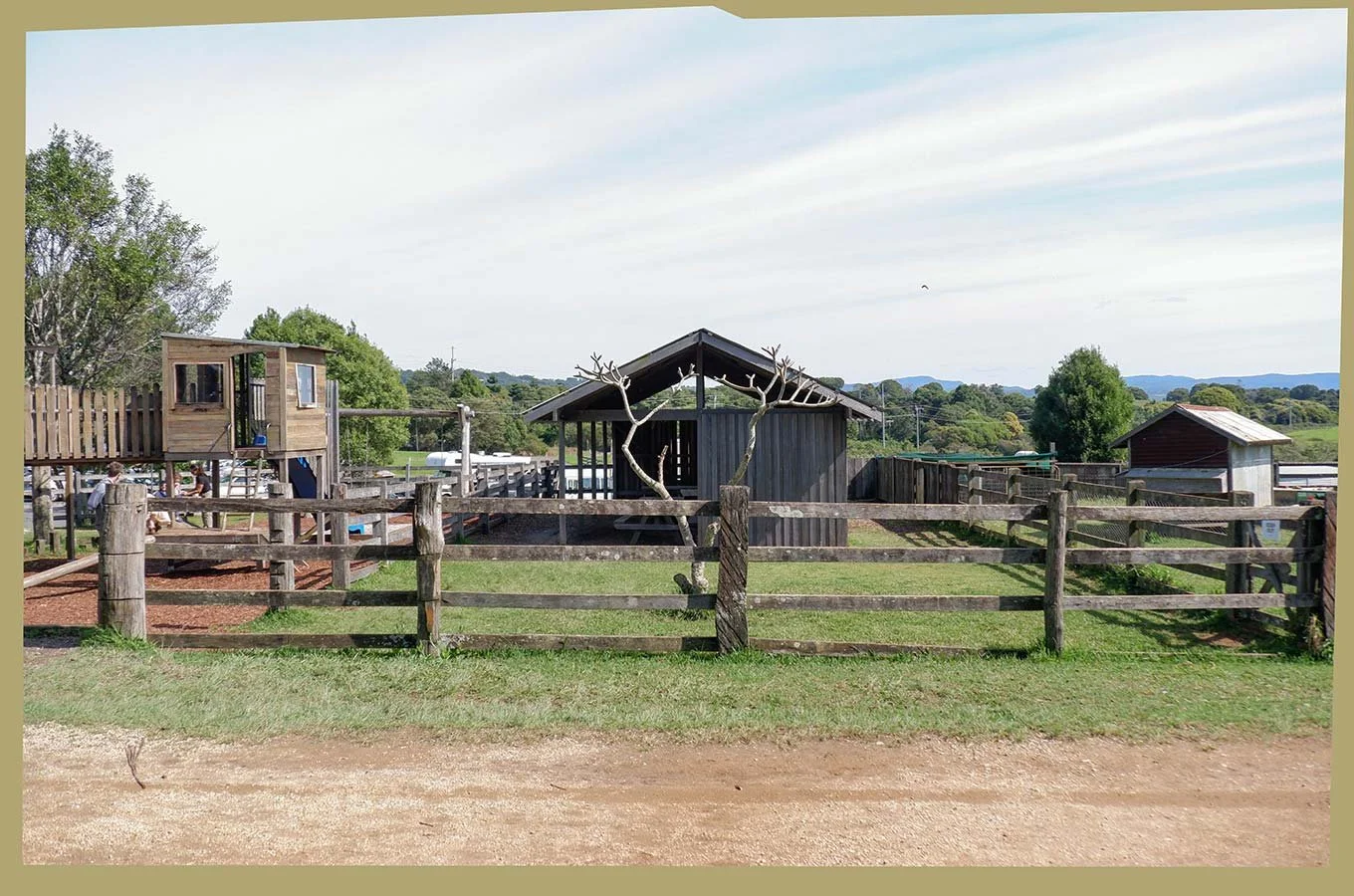 A rustic farm scene with a wooden fence, a tree with no leaves, and a wooden barn. There is a small wooden playhouse and a slide on the left side, with a few children playing nearby. The background features green fields, trees, and distant hills under a partly cloudy sky.