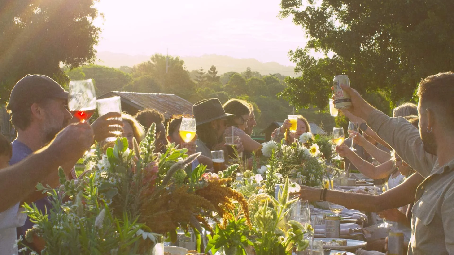 People gathered outdoors at sunset, raising glasses of wine and beer in a toast, surrounded by flowers and trees.