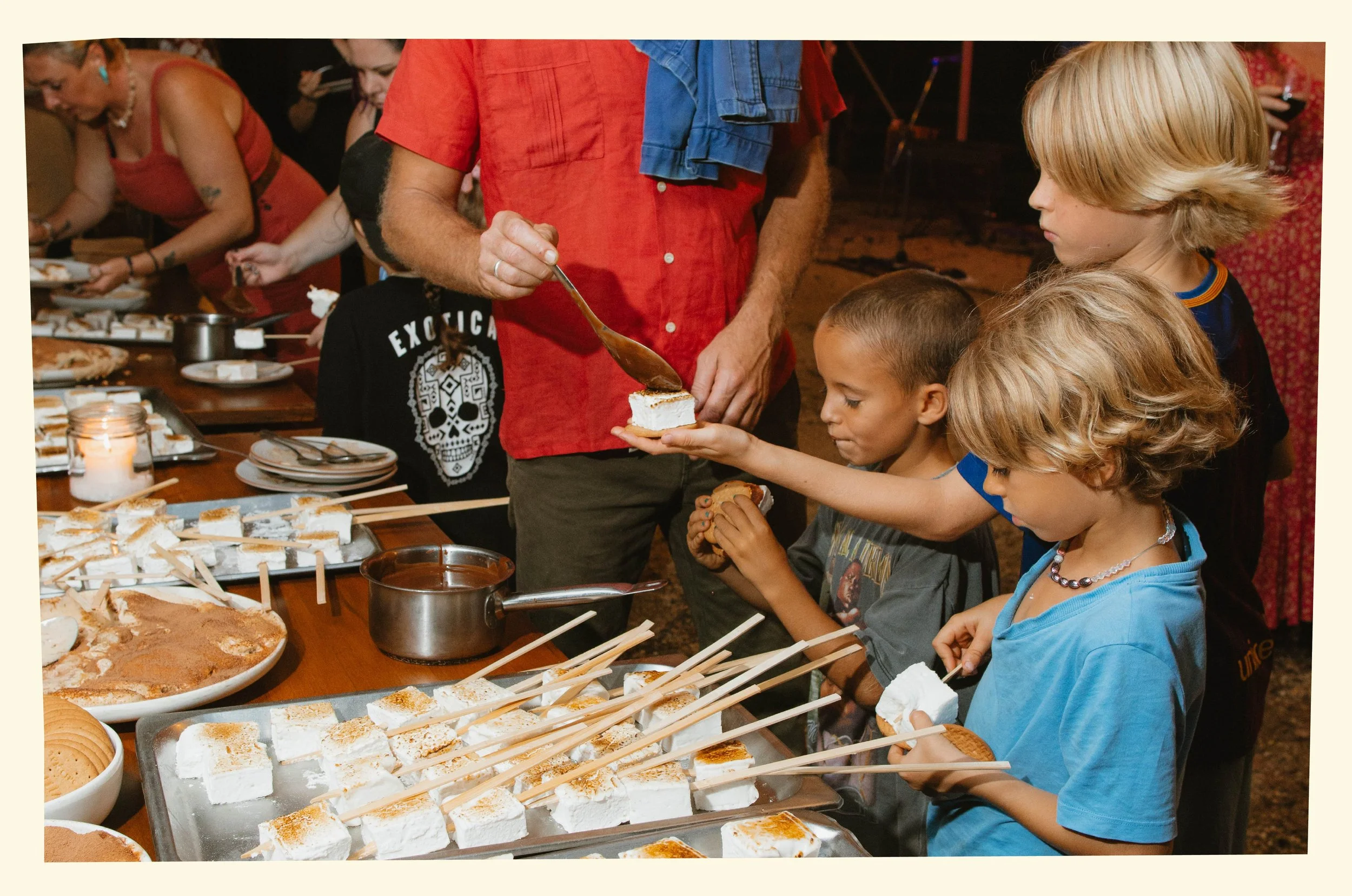 Children and adults at a dessert buffet, serving themselves marshmallows, cake, and other sweets at an indoor or outdoor gathering.