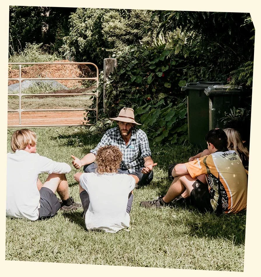 A man with a large wide-brimmed hat and beard sitting on the grass, talking to a group of children sitting around him outdoors in a grassy area near a fence and bushes.
