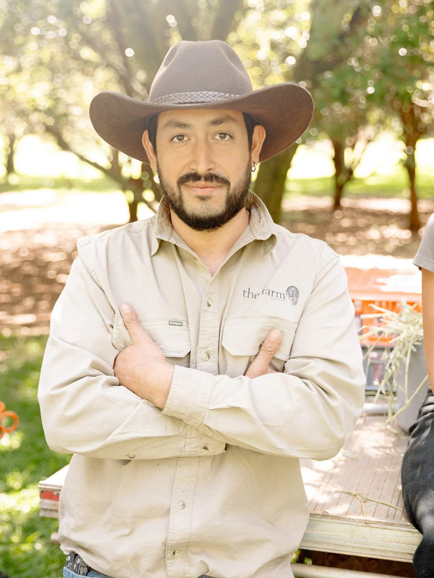 A man with a beard and mustache wearing a wide-brimmed cowboy hat and a light-colored shirt with a logo on the chest, standing outdoors with trees and sunlight in the background.