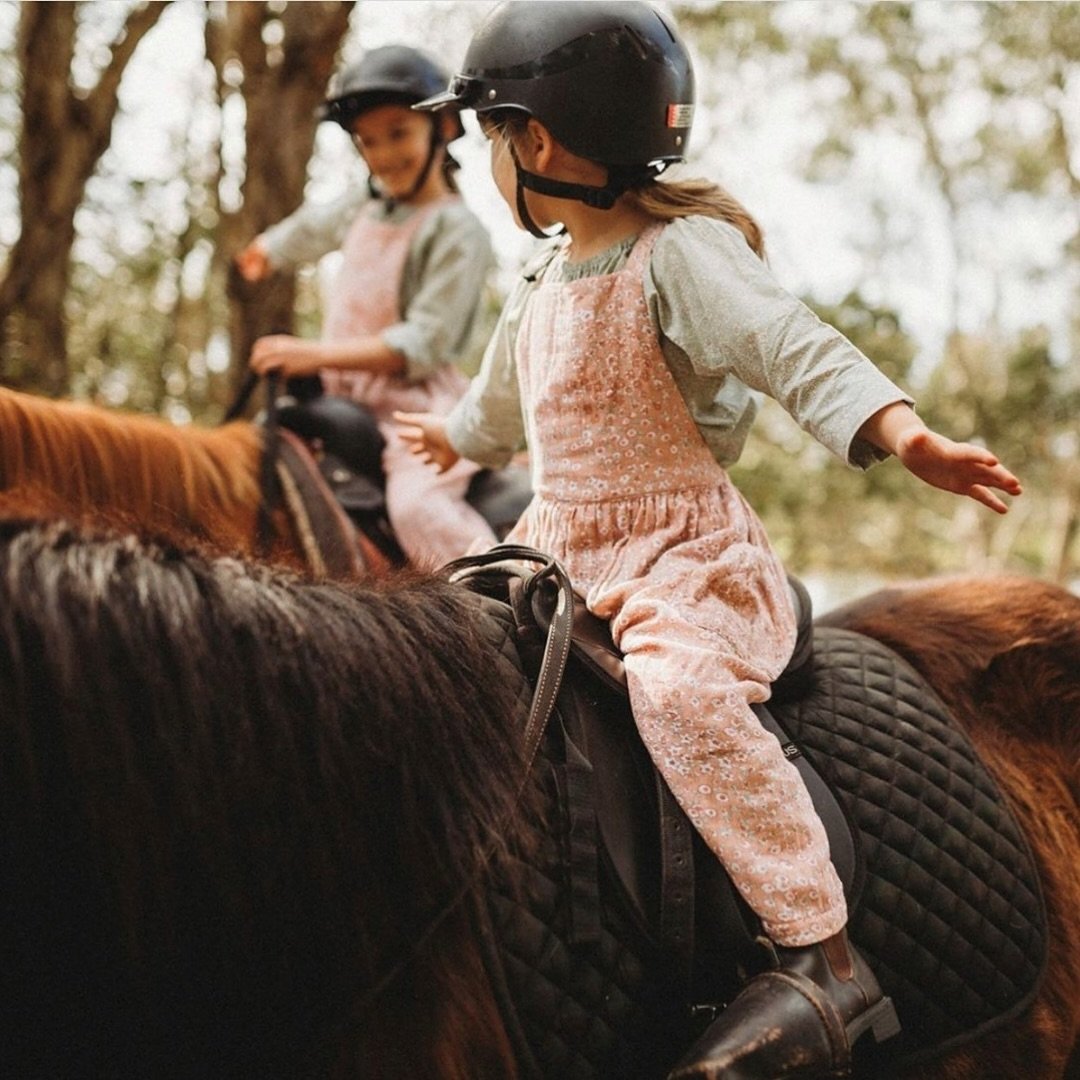 Two young girls riding horses outdoors, wearing helmets and casual clothing, with trees in the background.