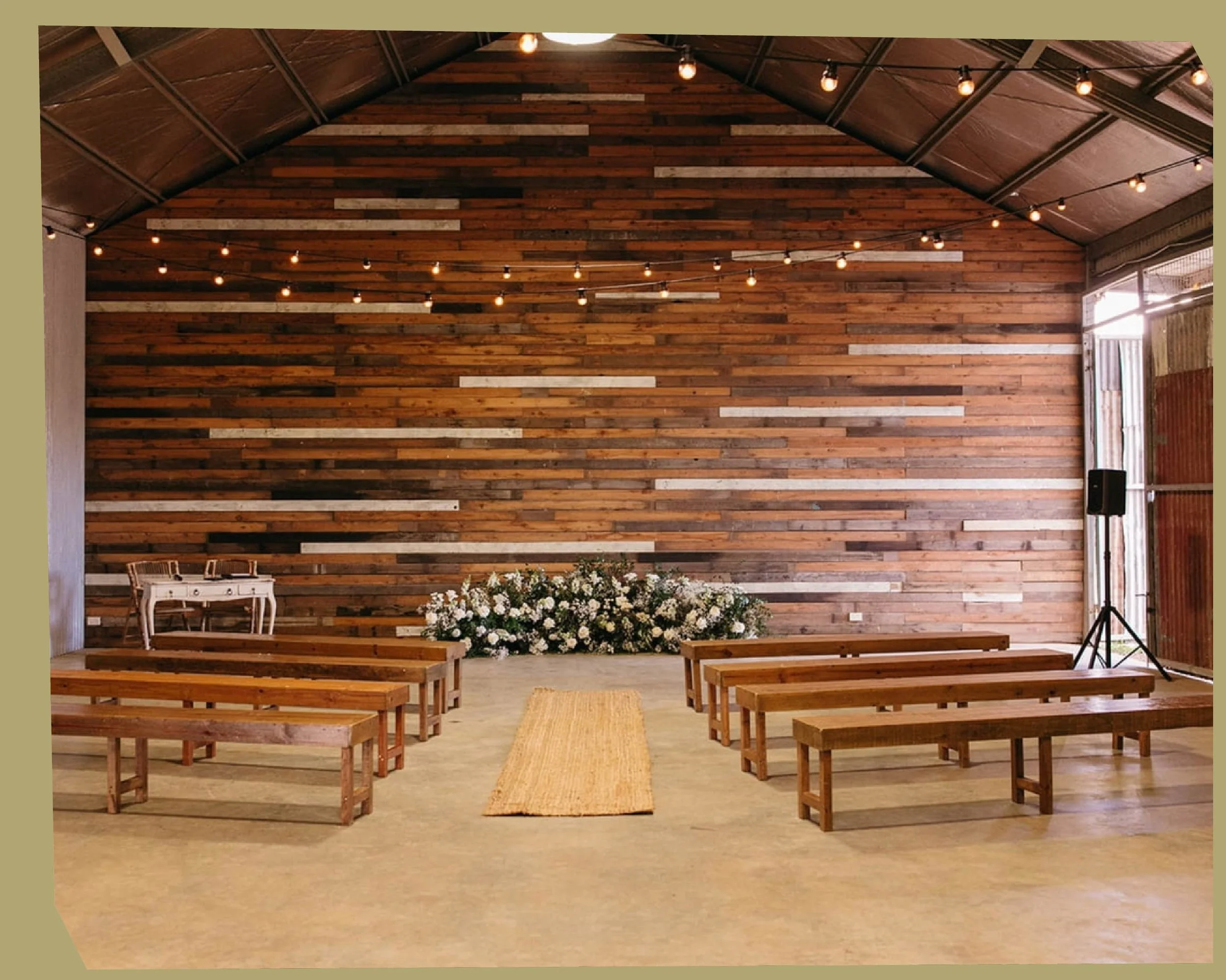 Interior of a rustic wooden event space with benches, a podium, a floral arrangement, and string lights hanging from the ceiling.