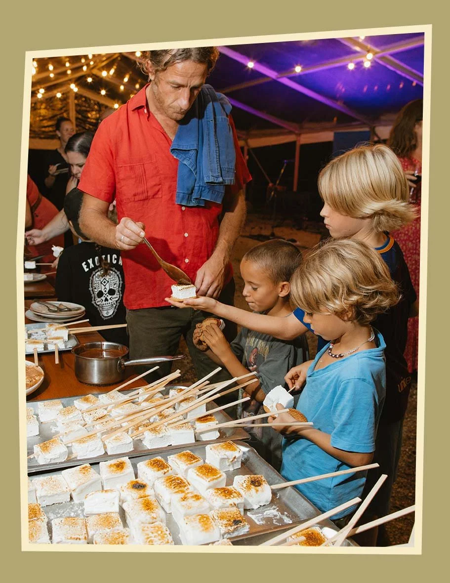 Children at a food stand selecting toasted marshmallows on sticks under a tent with string lights