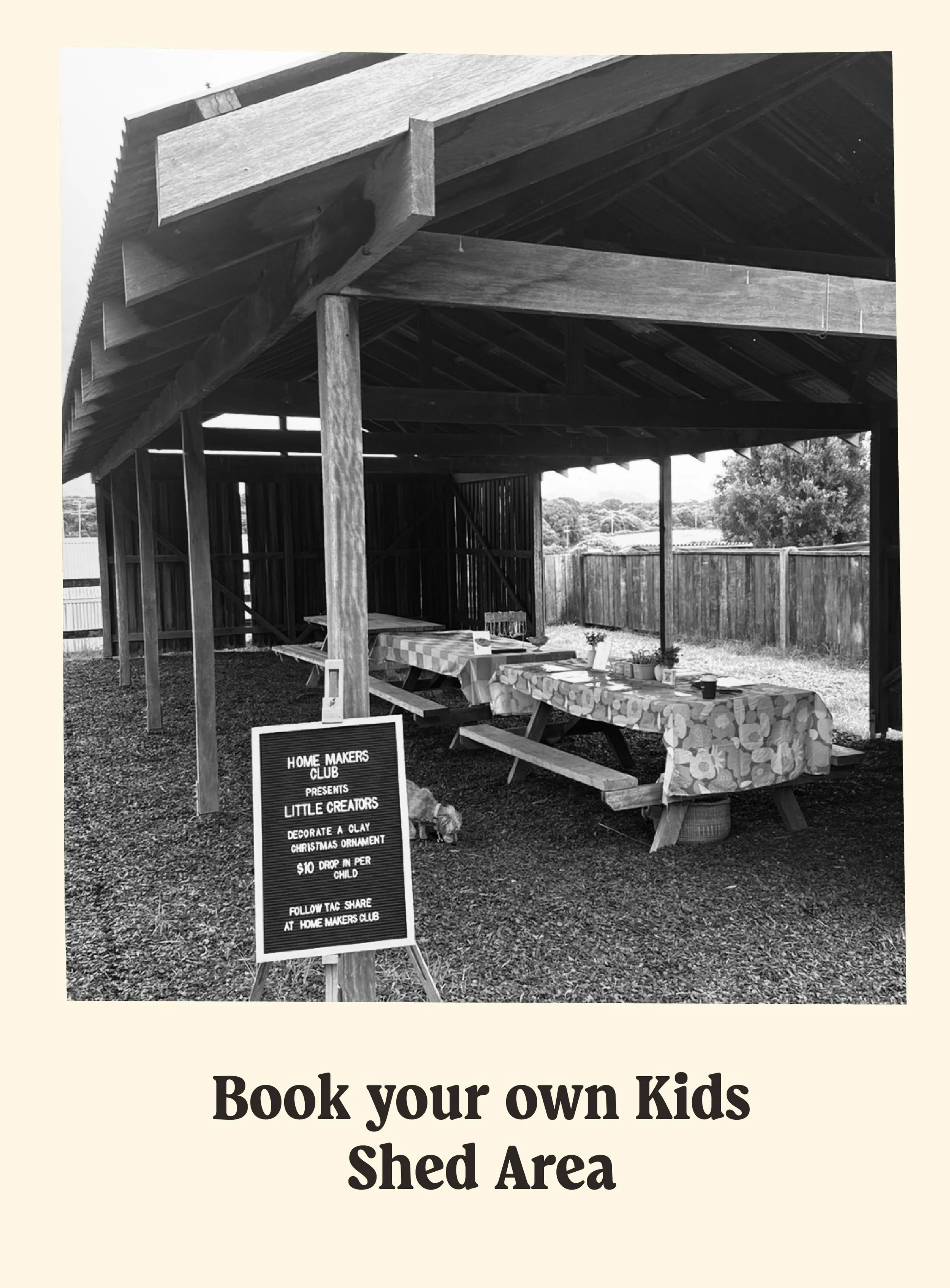 Black and white photo of a wooden outdoor shed with tables inside for kids activities, set up for an event. Signboard advertises a kids craft activity, decorating a clay Christmas ornament for $10, at a Kids Shed Area.