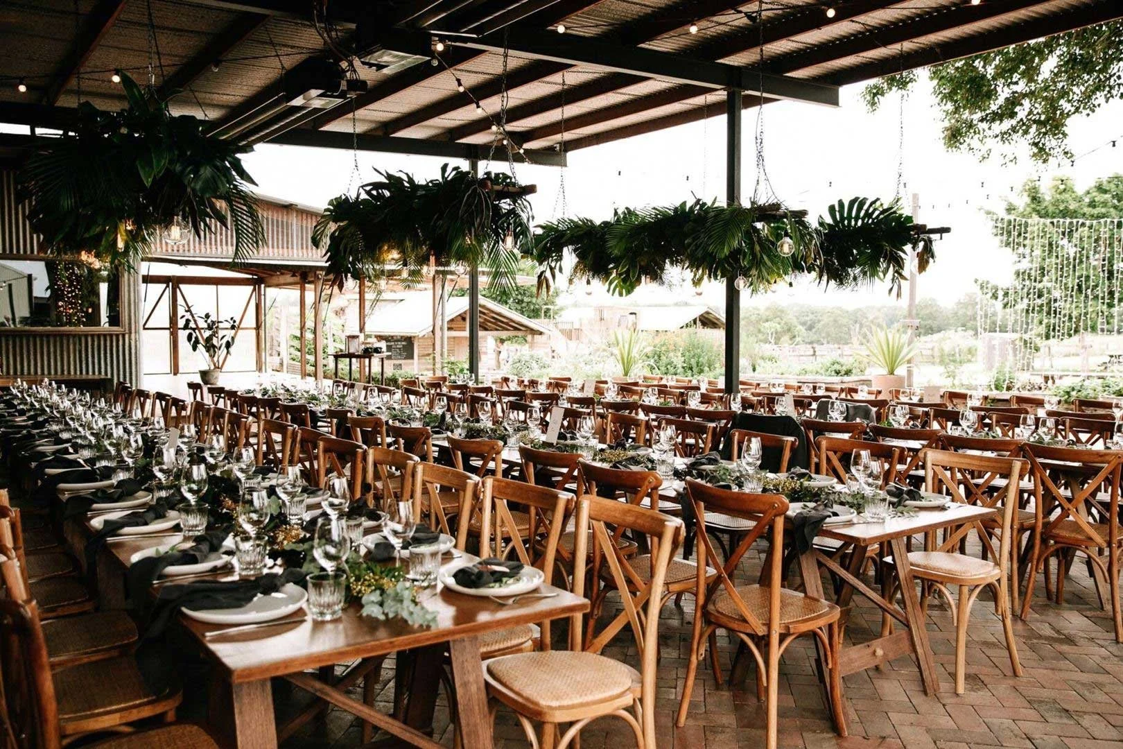 A large outdoor event space with long wooden tables set for a formal gathering, decorated with greenery and black napkins, under a covered roof with hanging plants and string lights.