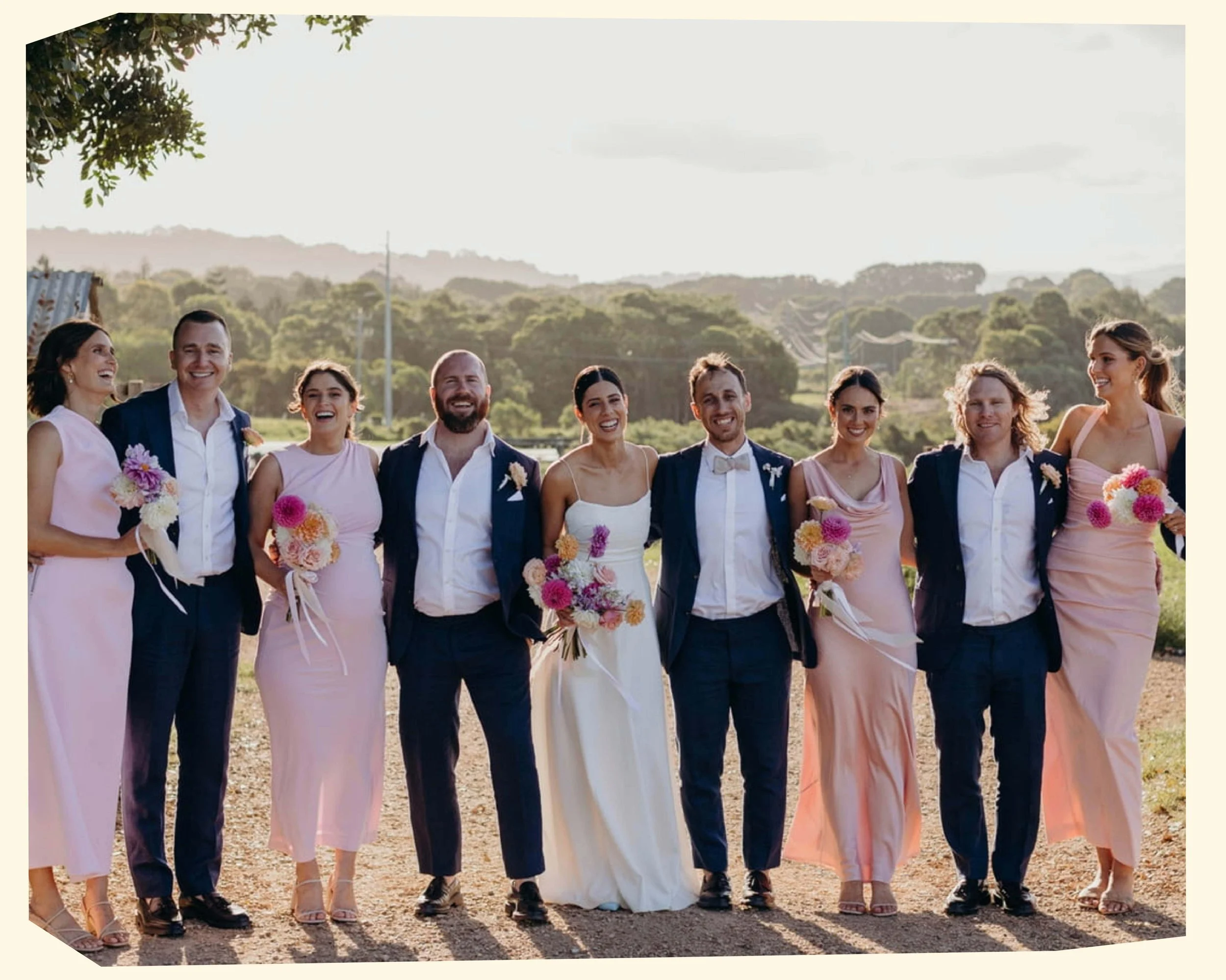 A group of nine people, including a bride and groom, standing outdoors with trees and hills in the background, smiling and holding flowers, dressed in wedding attire.