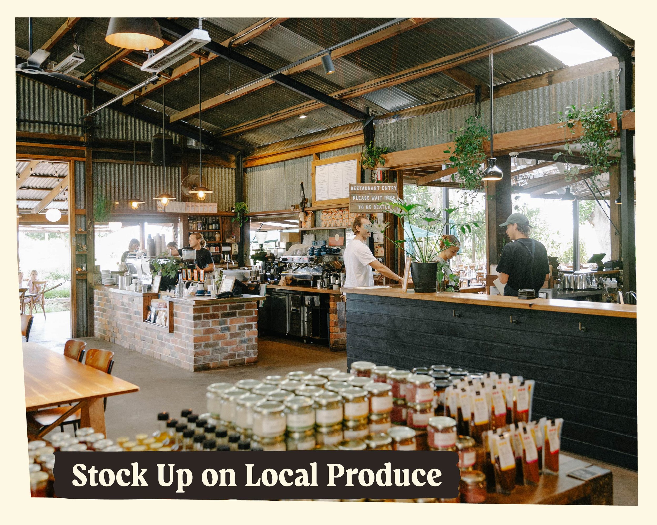 Interior of a rustic cafe or store with a brick and wood counter, shelves with products, and a sign that says 'Stock Up on Local Produce.' There are customers and staff inside, with natural light coming from large windows.