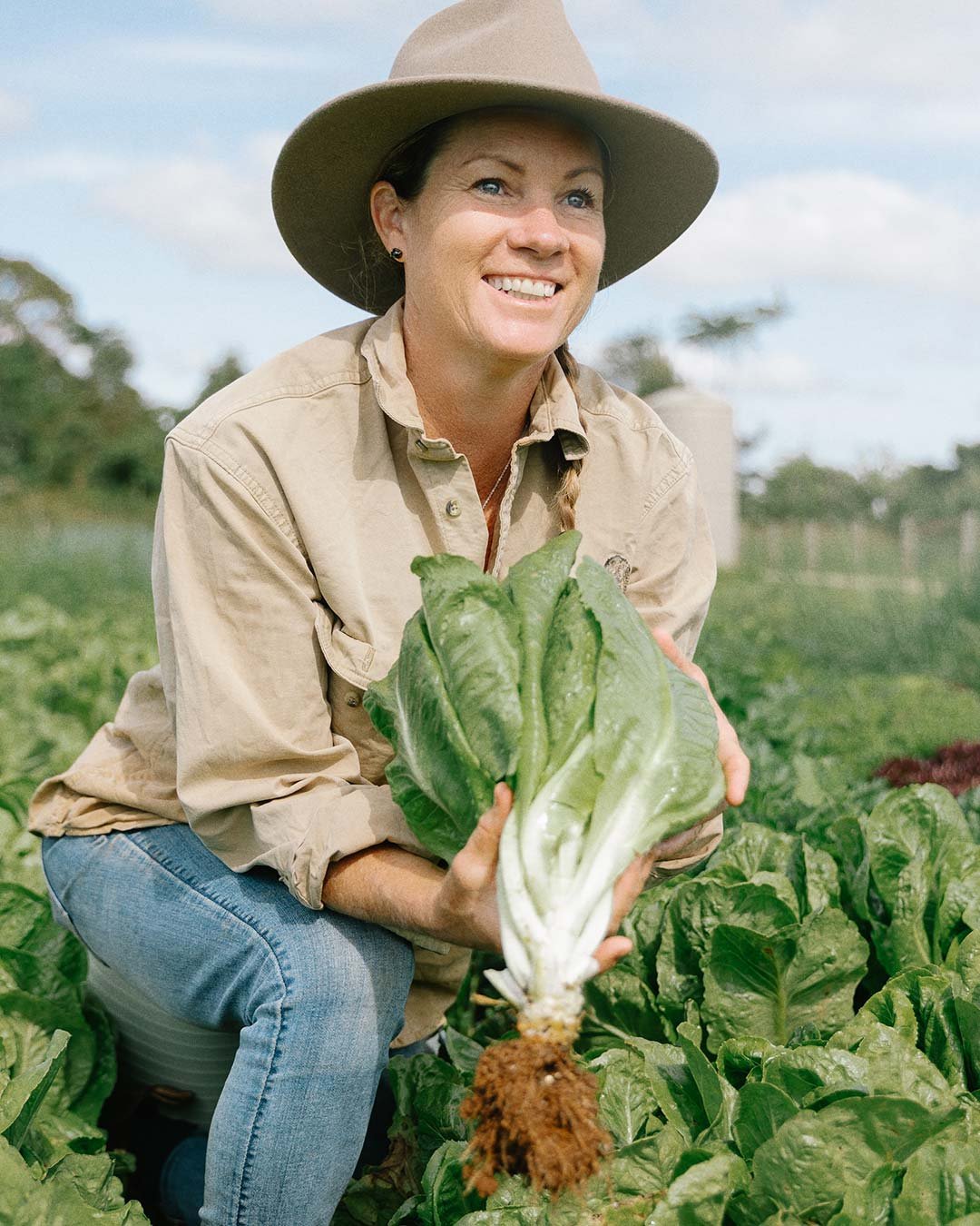A woman in a beige long-sleeve shirt and wide-brimmed hat kneels in a field of lettuce, holding a large head of lettuce with a smile on her face.