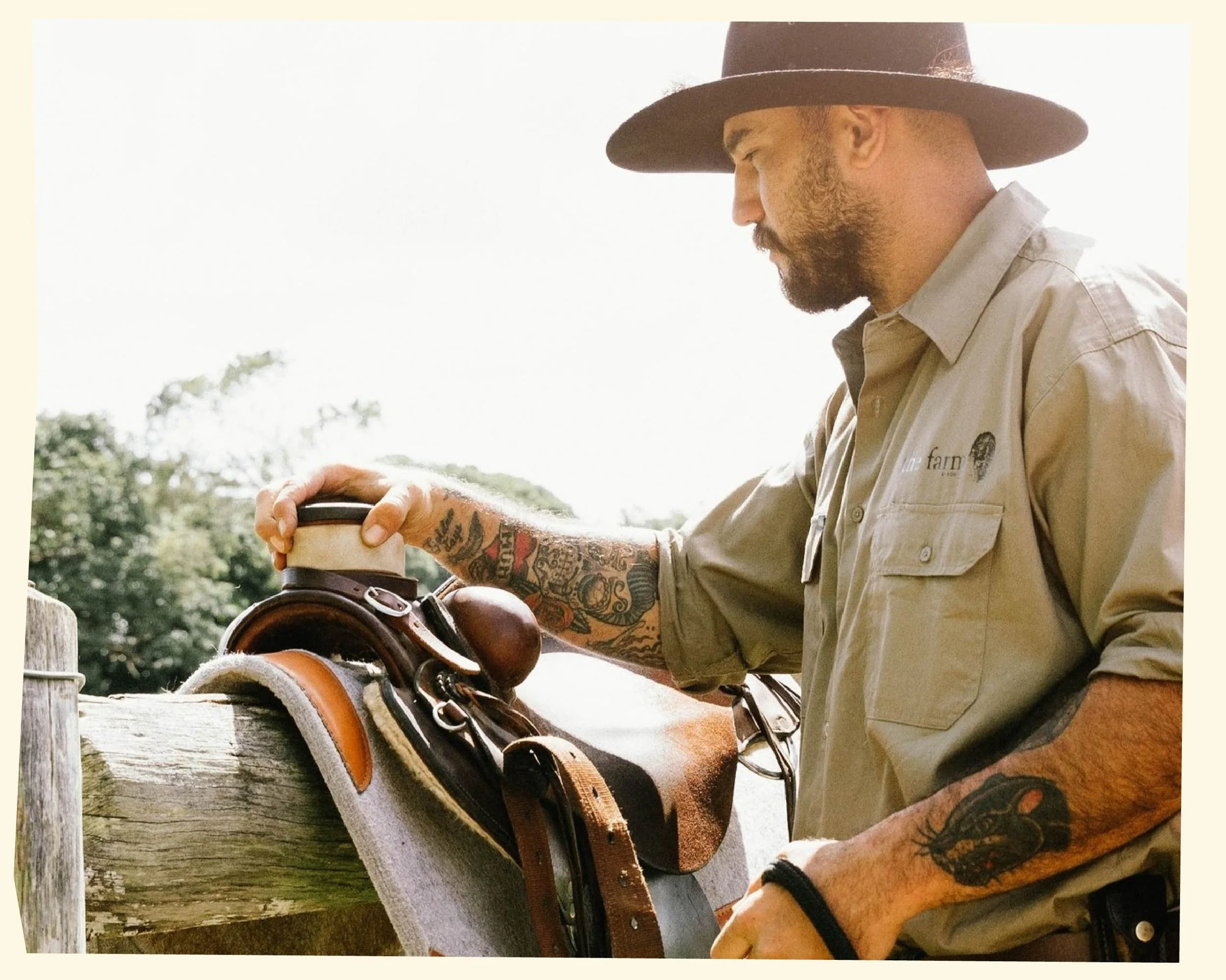 A man with tattoos wearing a wide-brimmed hat and beige shirt, standing outdoors near a wooden fence with trees in the background, holding a cup and preparing to ride a horse with a saddle and bridle.