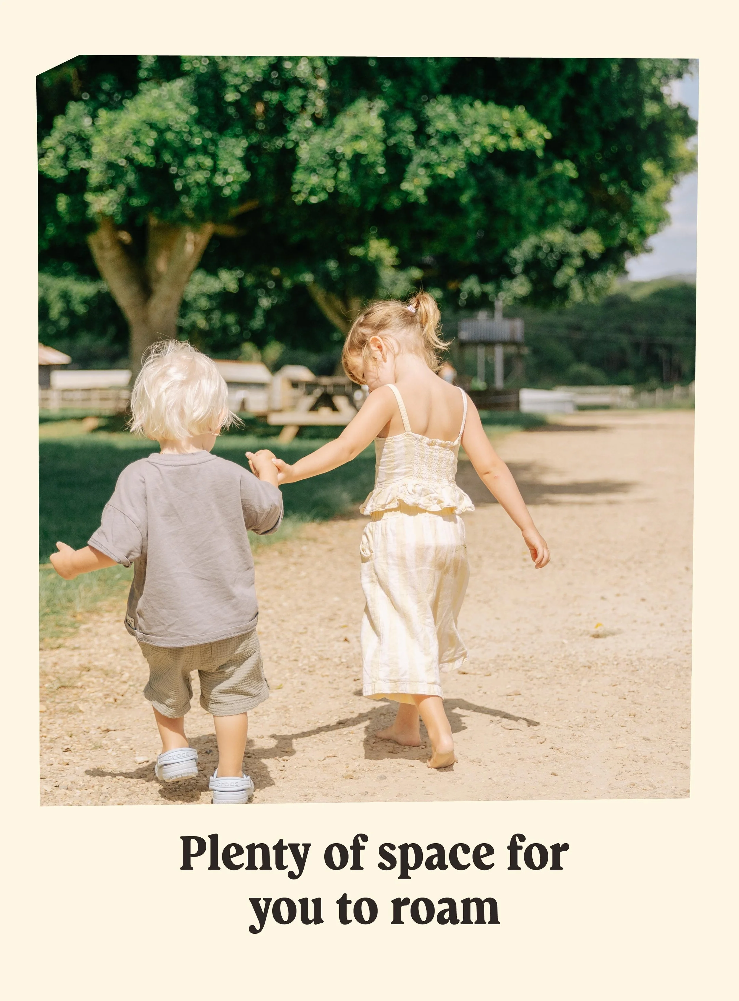 Two young children, a girl and a boy, walking barefoot on a dirt path in a park, holding hands. The girl is wearing a light-colored dress, and the boy is wearing a gray t-shirt and shorts. There are large trees and picnic tables in the background.
