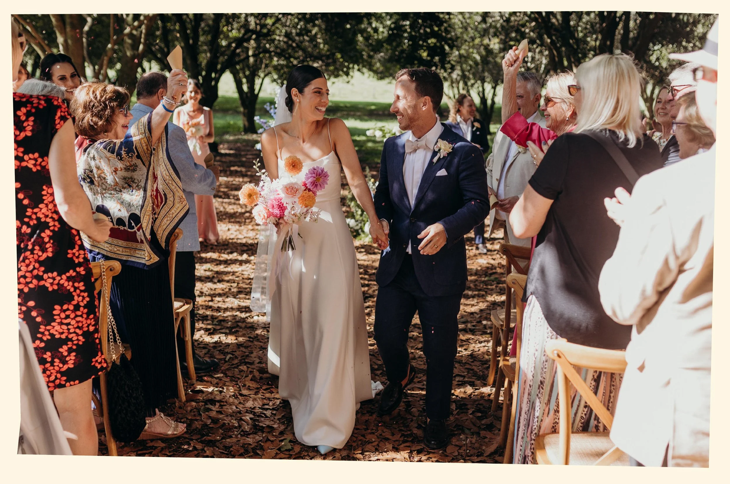 A bride and groom holding hands at an outdoor wedding ceremony surrounded by smiling guests, trees, and sunlight.