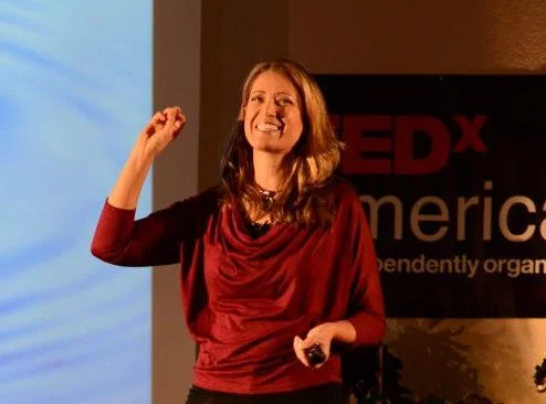 Woman smiling and gesturing with her right hand, standing in front of a TEDx banner.