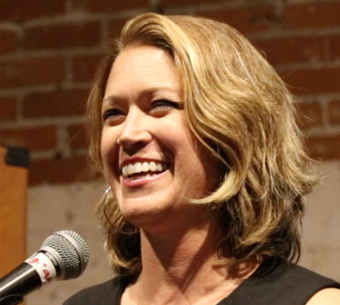 Dr. Jenn Gunsaullus with shoulder-length wavy blonde hair smiling while speaking into a microphone, with a brick wall background, speaking about consent, sex, and gender topics.