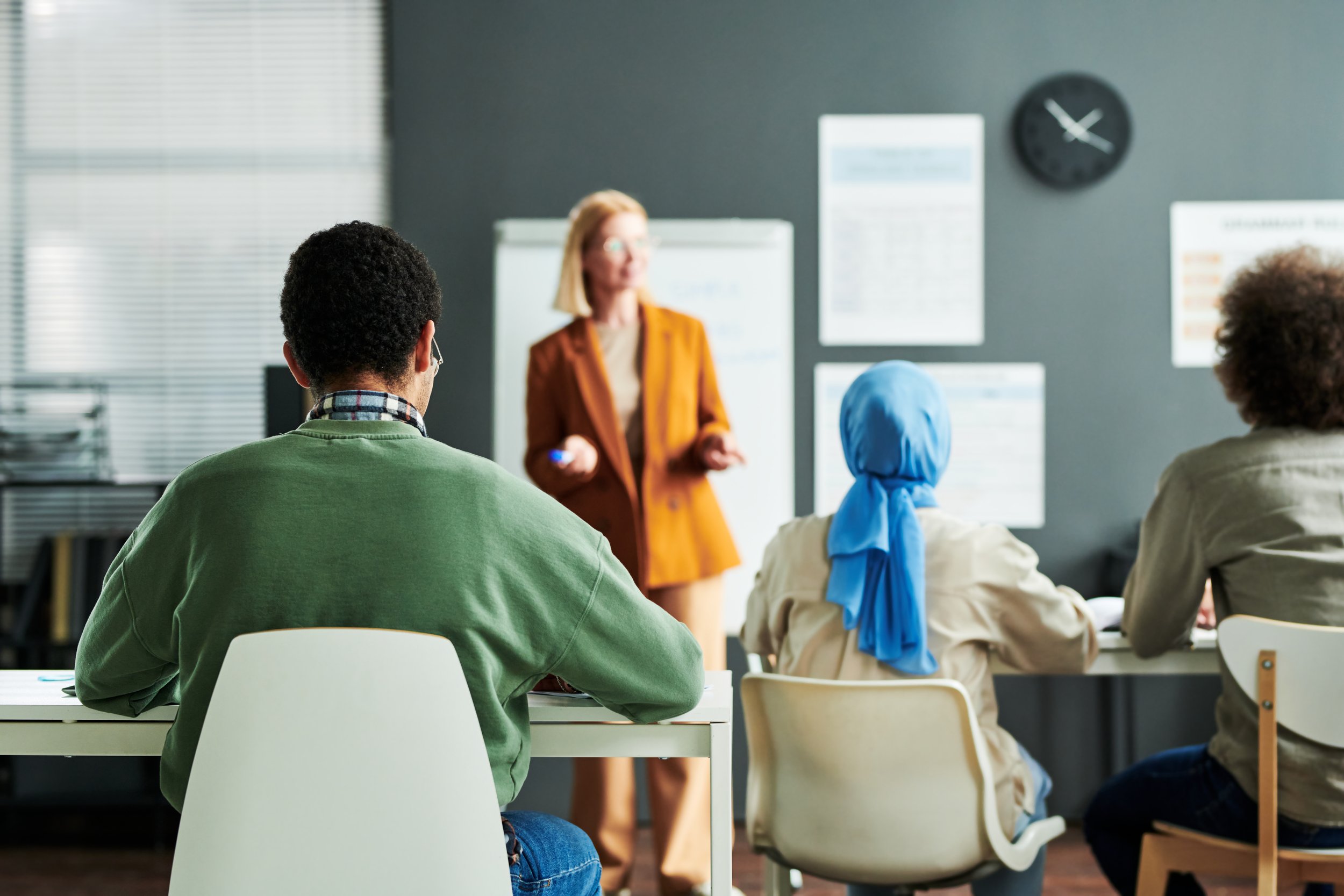 A diverse group of people attending a classroom or workshop, listening to a woman instructor who is speaking in front of a dark wall with charts and a clock.