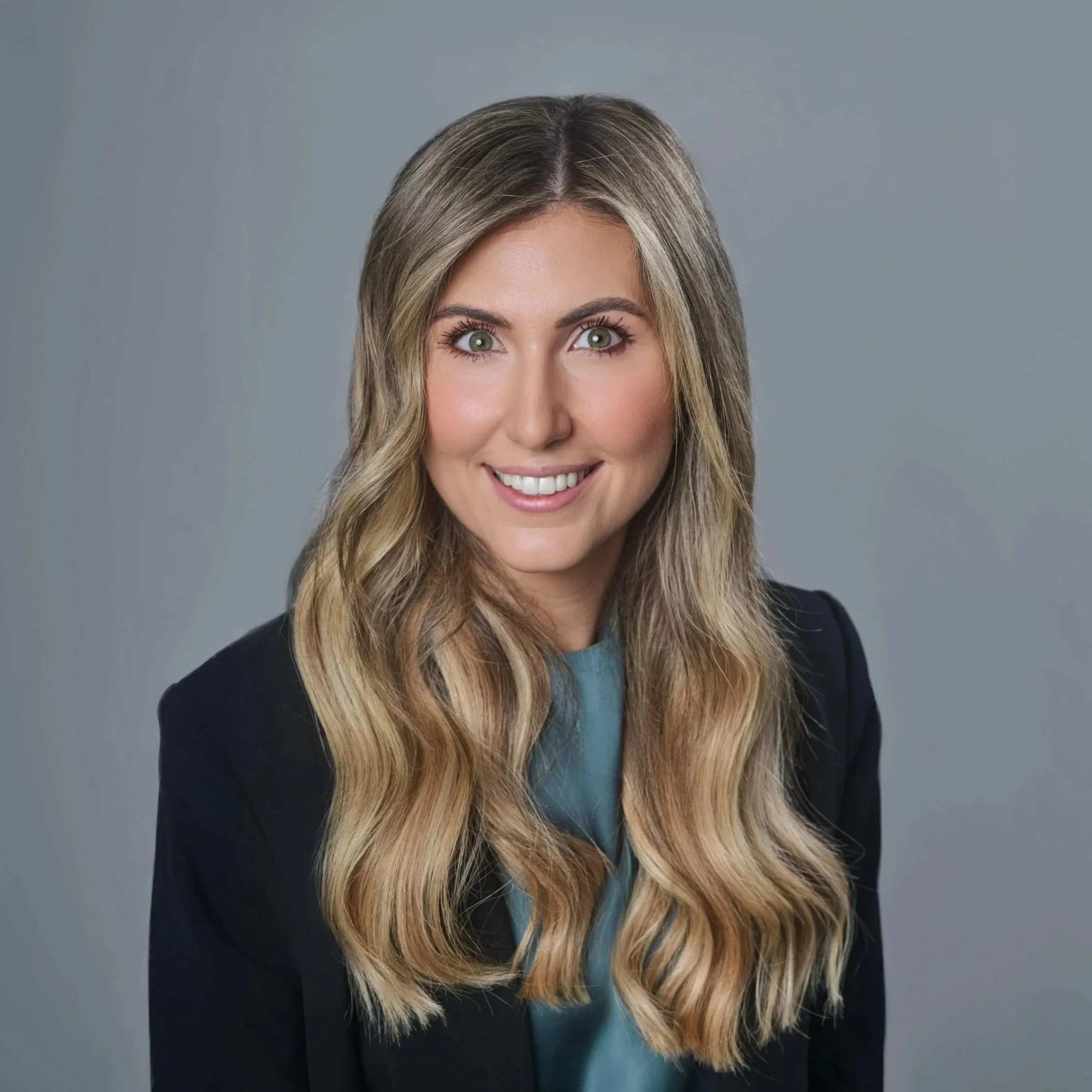 A woman with long, wavy blonde hair smiling, wearing a dark blazer and a teal blouse against a gray background.